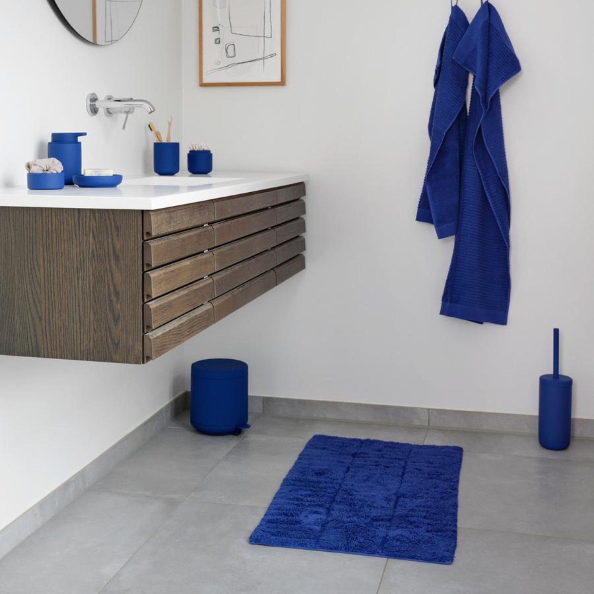 Moderne badkamer met een zwevend houten badmeubel, witte wastafel, blauwe accessoires uit de Ume collectie, een blauwe badmat, Zone Denmark Ume Toiletborstel Indigo Blauw en blauwe badjassen aan de muur. Lichtgrijze tegelvloer en minimalistische inrichting.