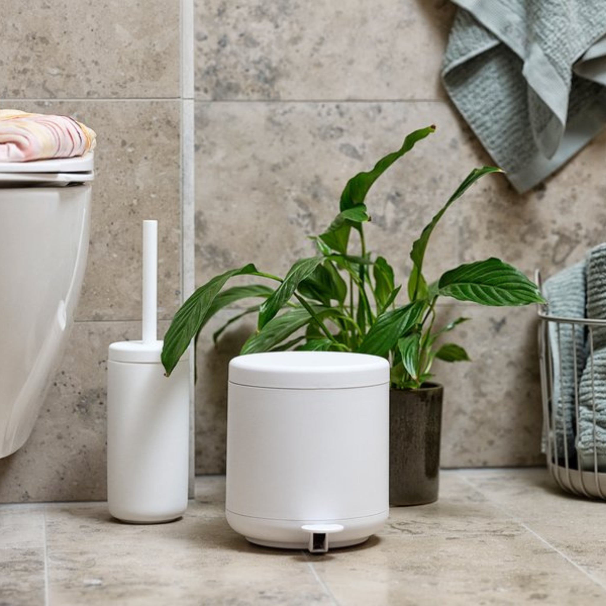 Een moderne badkamerscène met de Zone Denmark Ume Toiletborstel Wit, een witte pedaalemmer en een groene plant in pot op de betegelde vloer; handdoeken hangen vlakbij en links is een deel van een toilet zichtbaar.