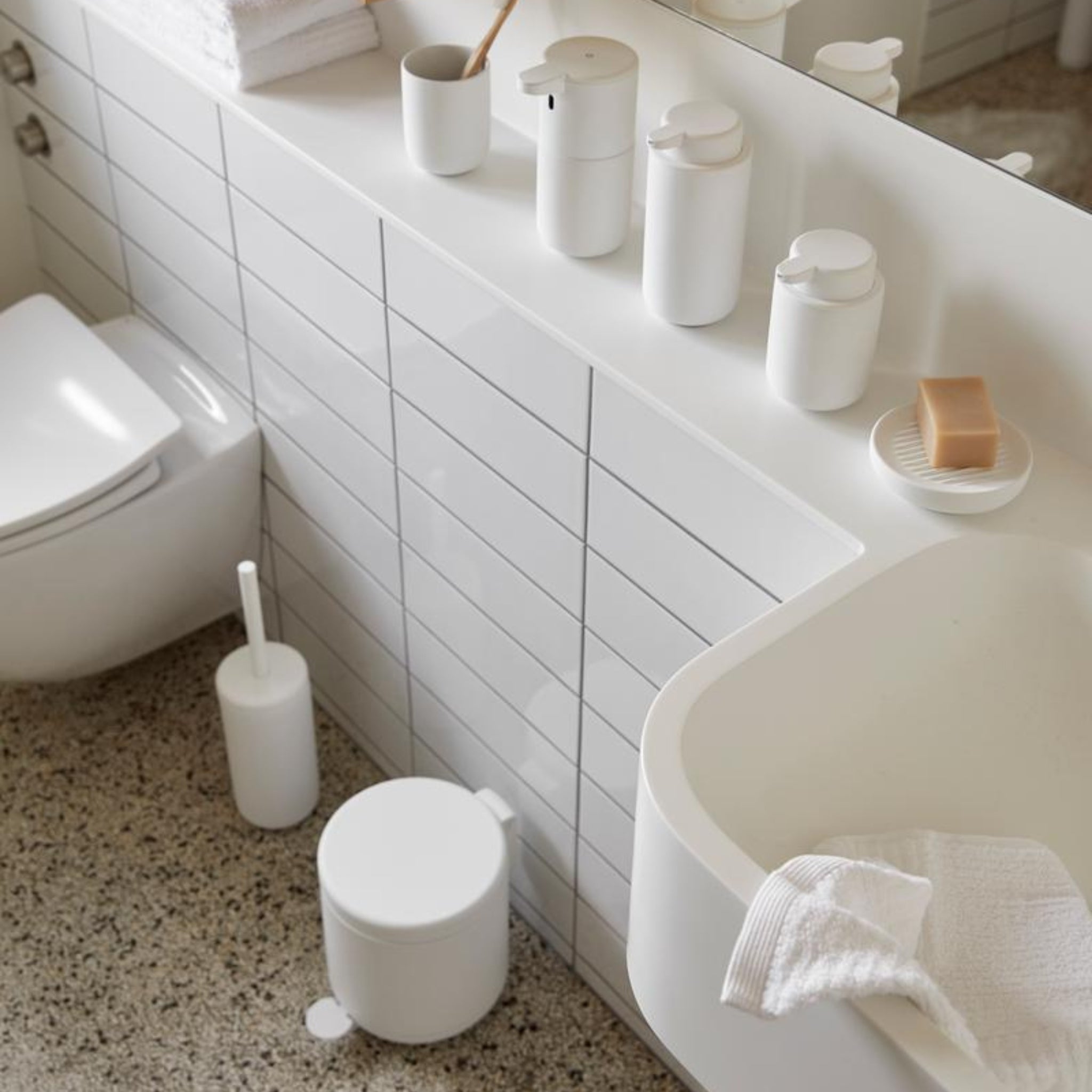 Een moderne, witte badkamer met de Zone Denmark Ume Toiletborstel Wit, bijpassende prullenbak, zeepdispenser, bakjes, tandenborstelbeker, zeep op een schaaltje en een handdoek op het aanrecht.