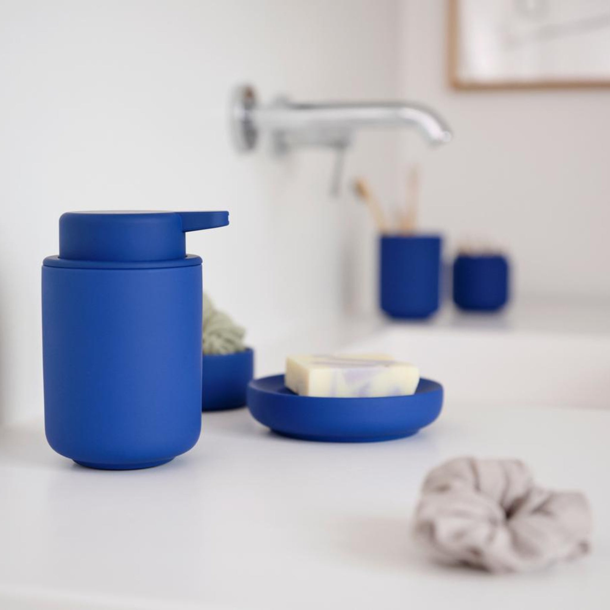 Een modern aanrecht met de Zone Denmark Ume Zeepdispenser in indigoblauw, bijpassend zeepbakje en beige scrunchie, met vage kraan en blauwe Zone Denmark accessoires op de achtergrond.