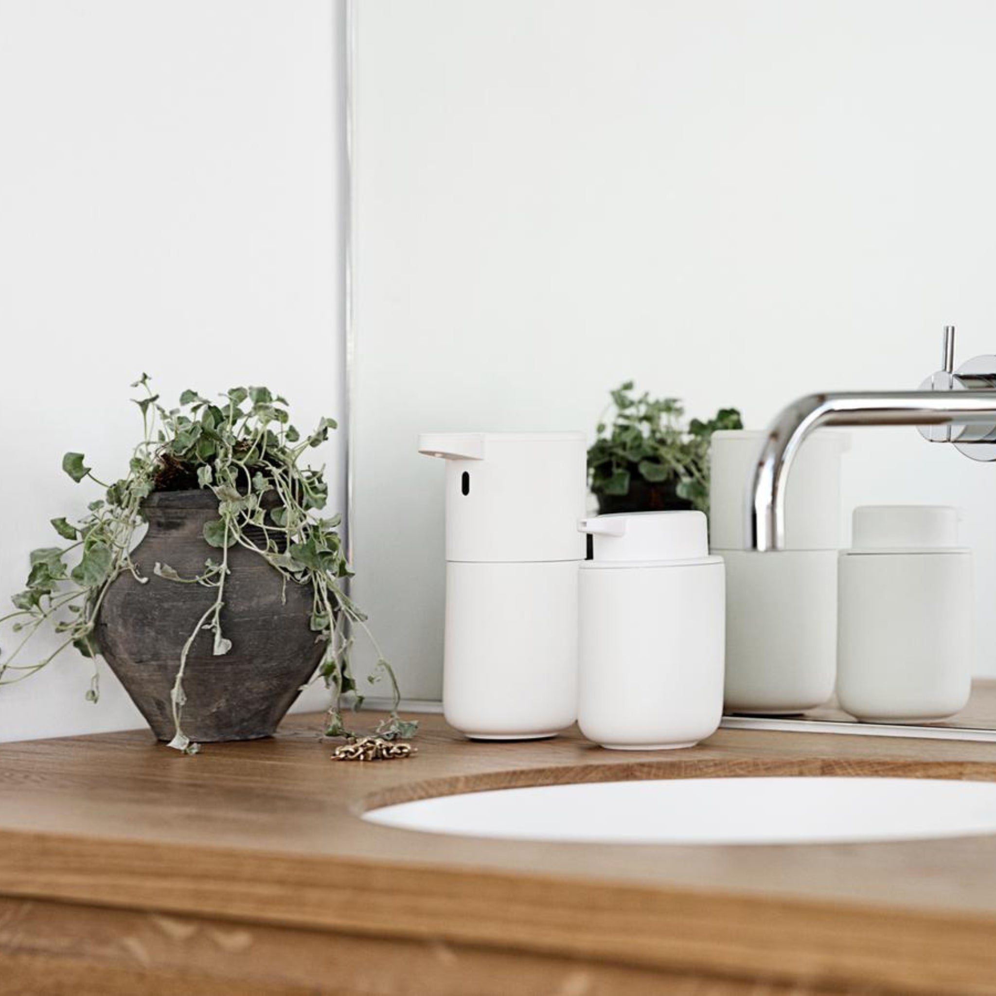 Een modern aanrecht met de Zone Denmark Ume Zeepdispenser Wit van Zone Denmark, samen met een potplant, kraan en spiegel boven een houten oppervlak met een witte inzetwastafel.