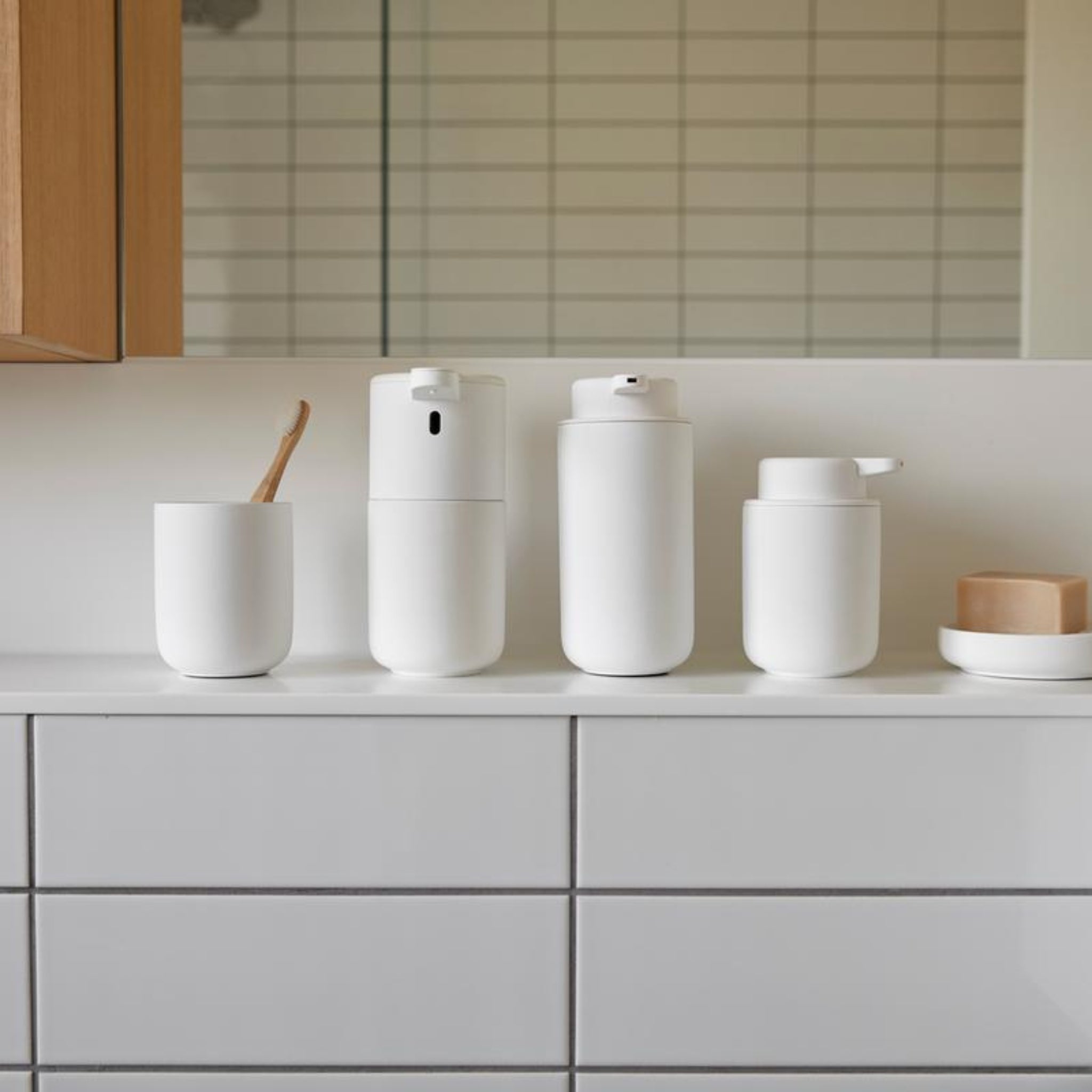 Een minimalistisch aanrecht bevat een witte tandenborstel in een beker, de Zone Denmark Ume Zeepdispenser Wit en een wit zeepbakje met zeep, allemaal gerangschikt bij een betegelde muur en spiegel voor een moderne badkamerlook.