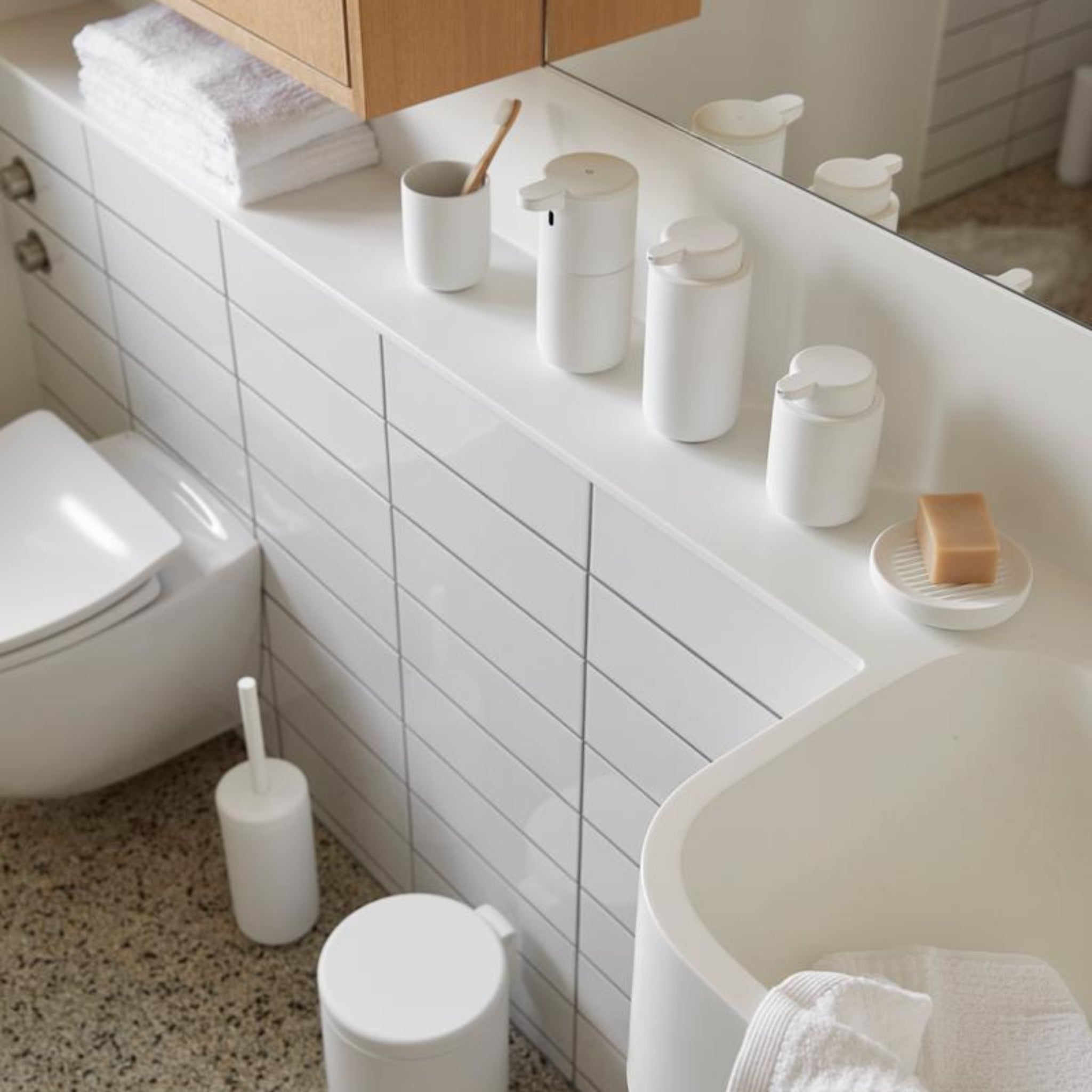 Moderne badkamer met witte tegels, accessoires zoals de Zone Denmark Ume Zeepdispenser Wit, een tandenborstelhouder, zeepbakje met zeep, gestapelde handdoeken, toilet, wc-borstel en kleine prullenbak op terrazzo vloer.