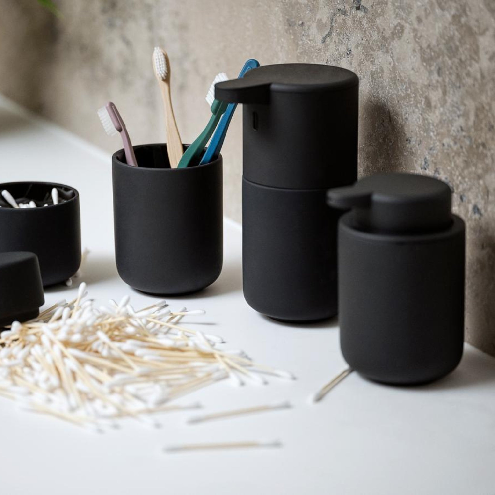 De Zone Denmark Ume Zeepdispenser Zwart, een bijpassende tandenborstelhouder met vier borstels en een wattenstaafjeshouder geven stijl aan het witte aanrecht in deze moderne badkamer, tegen een gespikkelde muur.