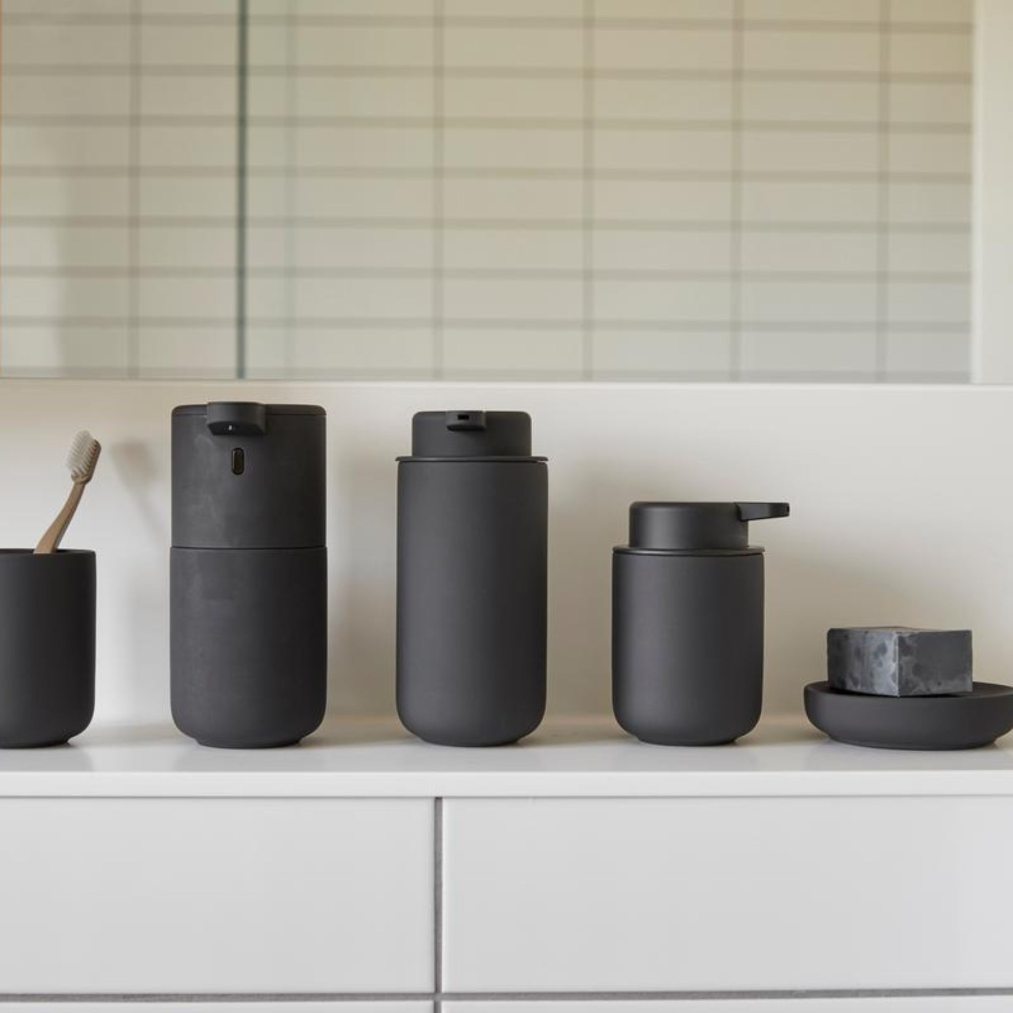 Een rij matzwarte Zone Denmark accessoires, waaronder de Ume Zeepdispenser Zwart, beker met tandenborstel, bus en zeepbakje met een rond stuk zeep, gerangschikt op een wit aanrecht in een moderne badkamer voor een betegelde muur.