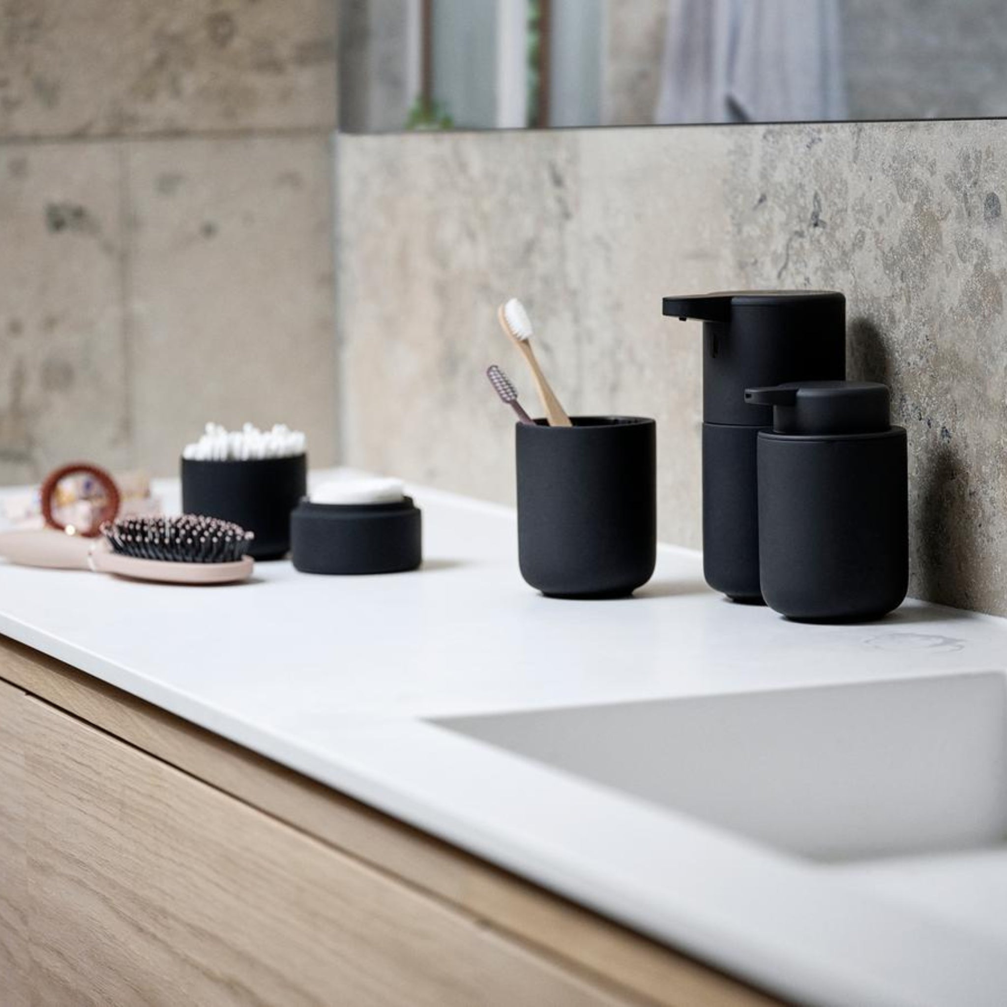 Een modern aanrecht met een witte wastafel, de Zone Denmark Ume Zeepdispenser Zwart, tandenborstelhouders met tandenborstels, een haarborstel, een potje wattenstaafjes en een ronde container tegen een stijlvolle stenen muur.