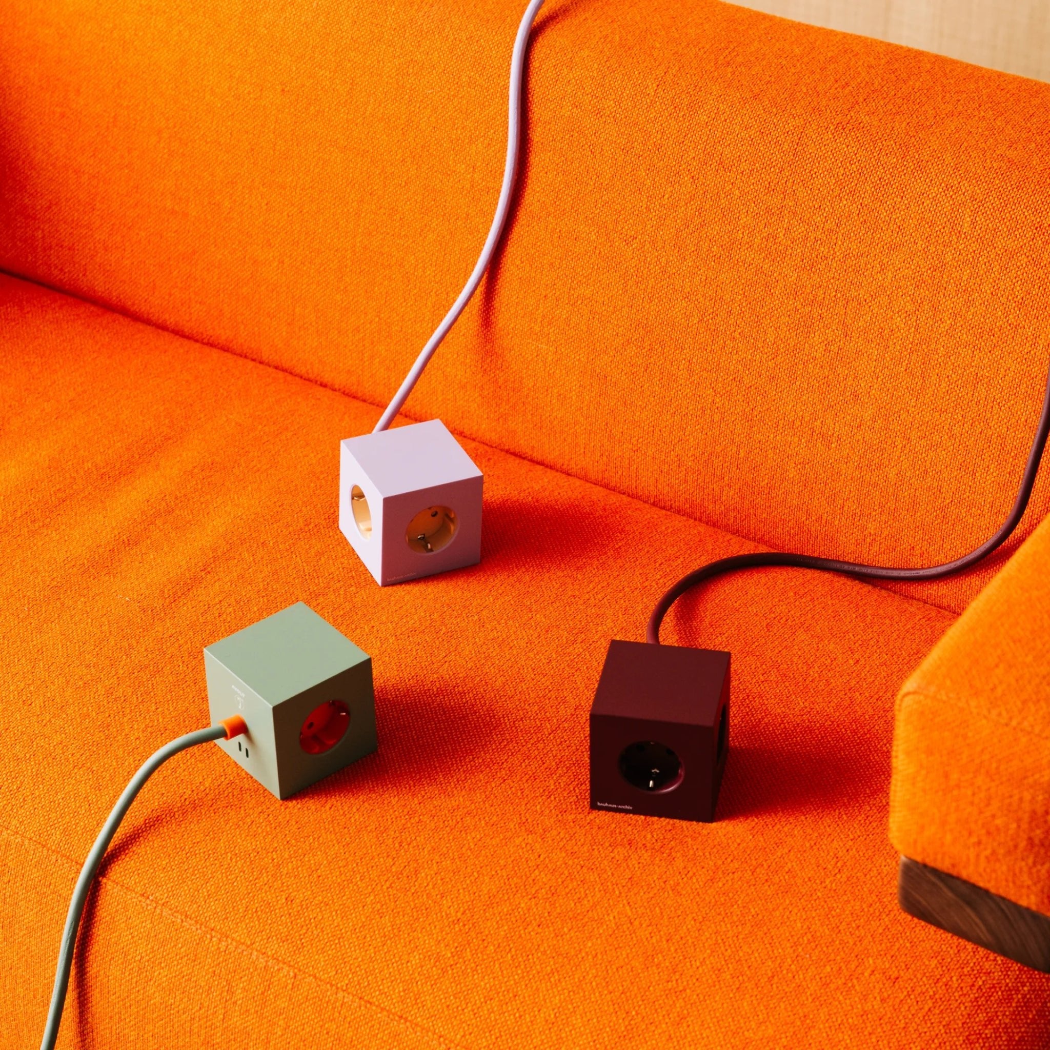 Drie Avolt x Bauhaus Archiv Square 1 USB-C Stekkerdoos stopcontacten in pastelgroen, lavendel en Mulberry Rood (1,8 m) voegen stijl toe aan een oranje bank, elk verbonden door een snoer in echt Bauhaus-design.