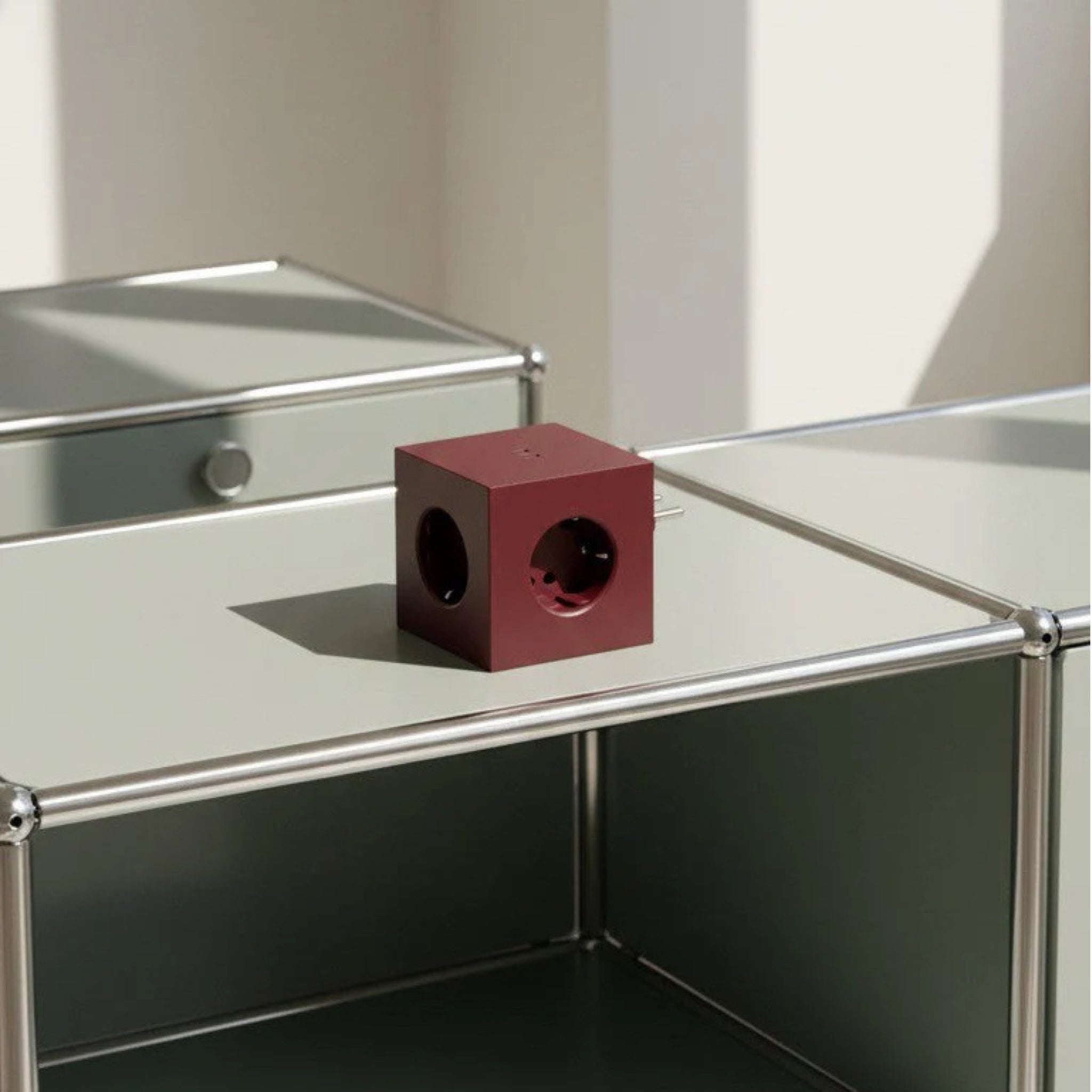 De Avolt x Bauhaus Archiv Square 2 USB-C Stekkerdoos Mulberry Rood van Avolt staat op een lichte metalen tafel met stalen buisranden, die een schaduw werpt in een heldere, minimalistische omgeving geïnspireerd op Bauhaus-design.