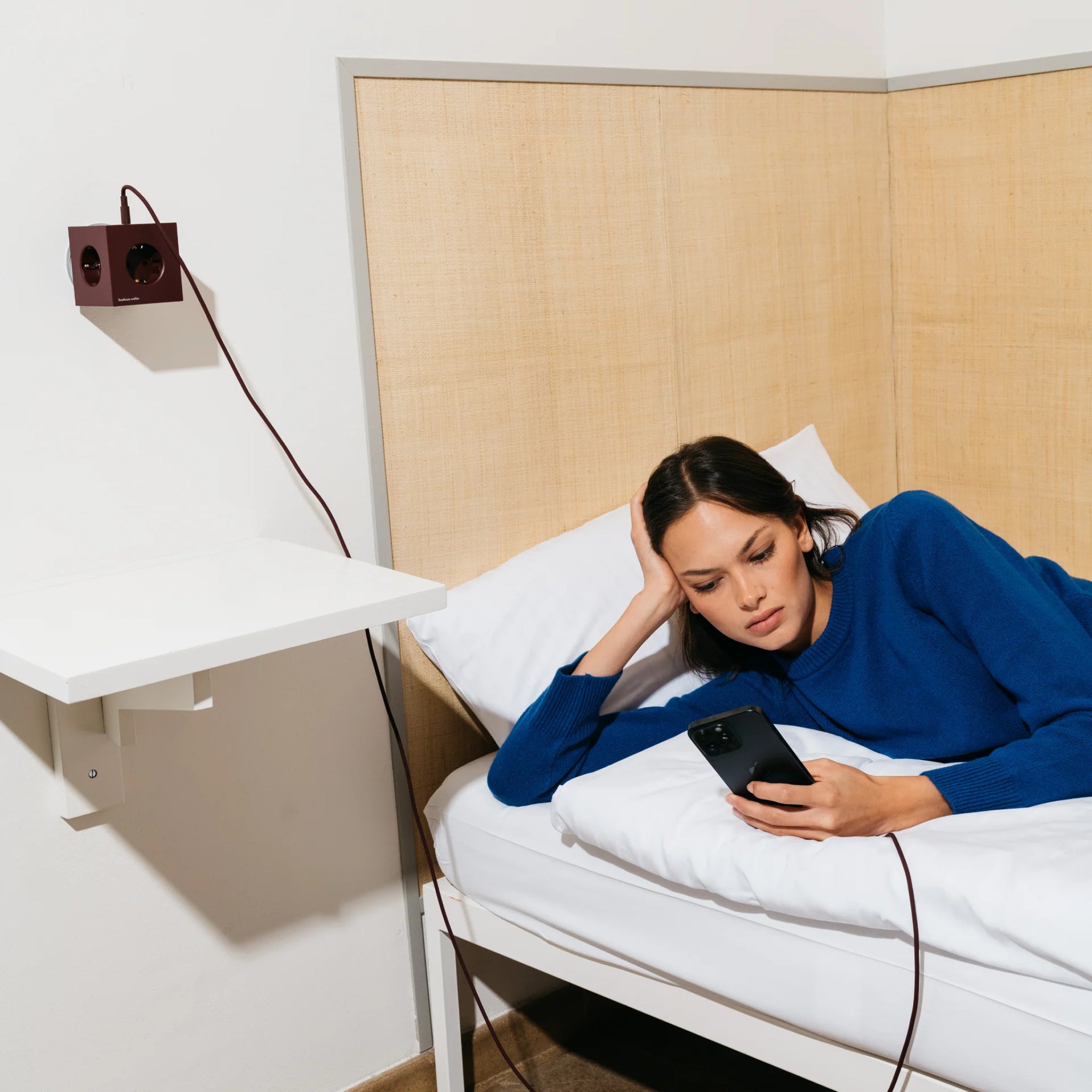 Een vrouw in een blauwe trui ligt op een bed en controleert haar telefoon die is aangesloten op een Avolt x Bauhaus Archiv Square 2 USB-C stekkerdoos Mulberry Rood, een stijlvolle wandgemonteerde optie die hoog bij een kleine witte plank is gemonteerd.