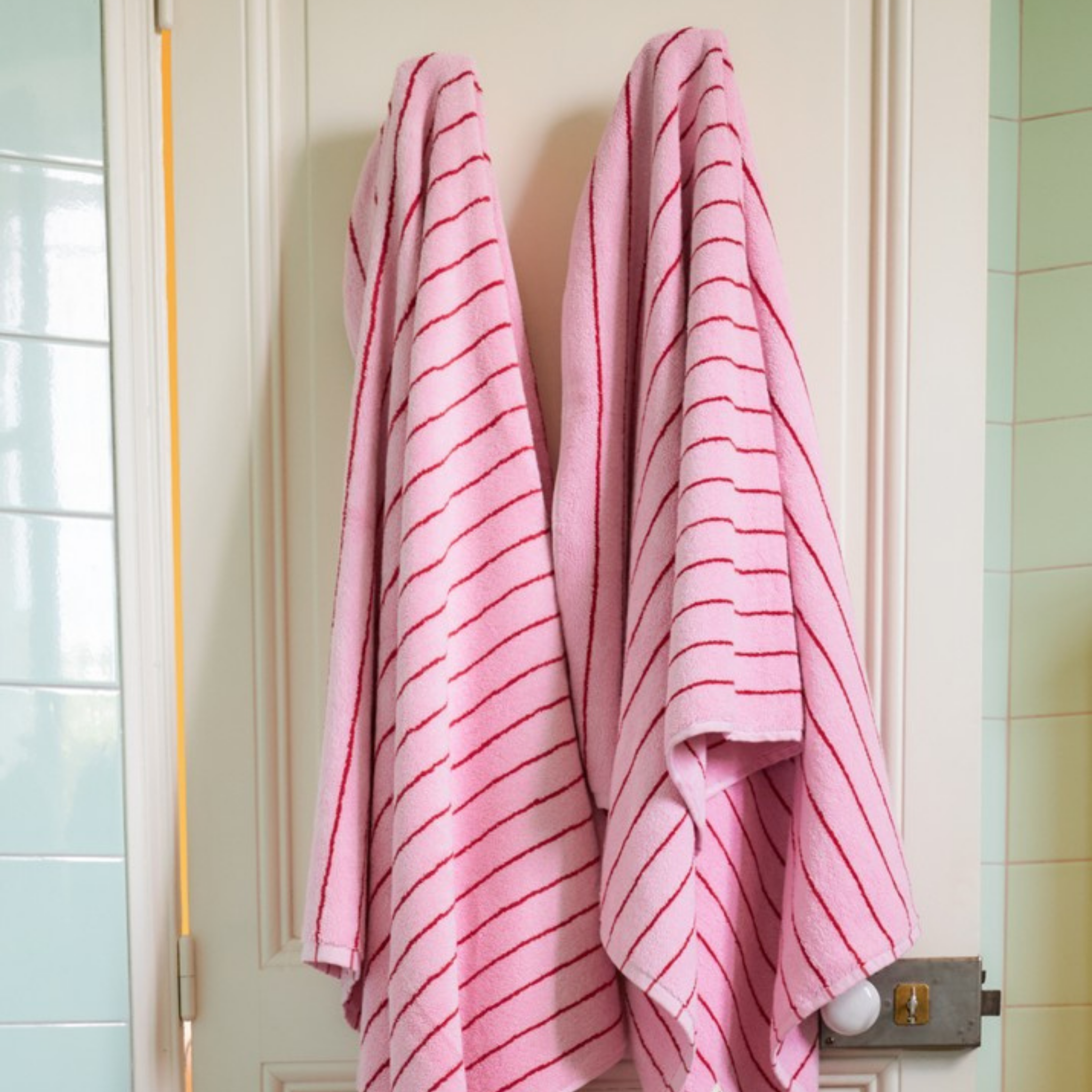Twee Bongusta Naram badhanddoeken in Baby Pink & Ski Patrol hangen zij aan zij op haken aan een witte deur in de badkamer. Op de achtergrond zie je lichtgroene en witte tegels.