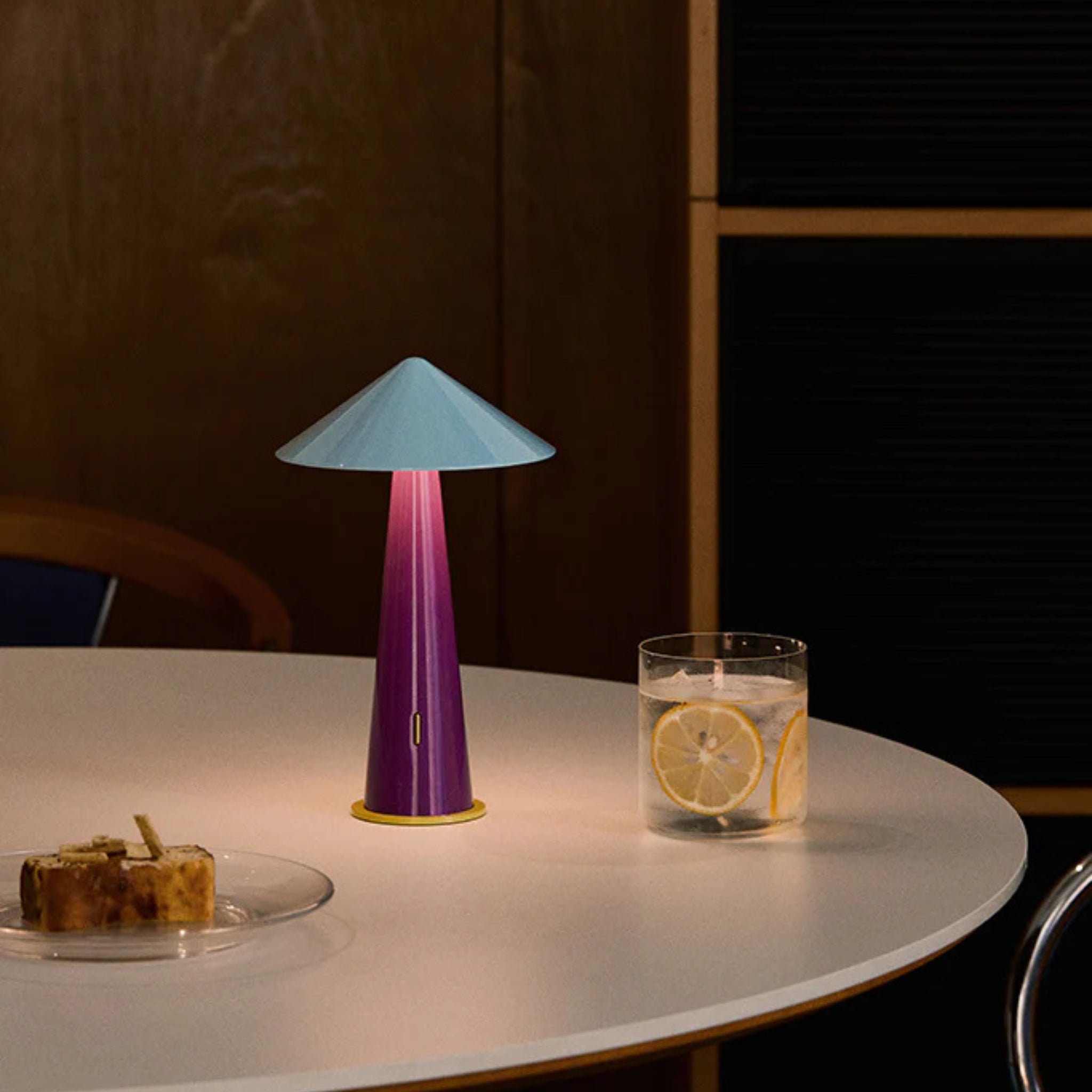 De Fine Lumens Tipsy Lamp LED Oplaadbaar Grijsblauw Pruim staat dimbaar op een ronde witte tafel naast een glas water met schijfjes citroen en gebak, tegen een houten en zwarte lambrisering.