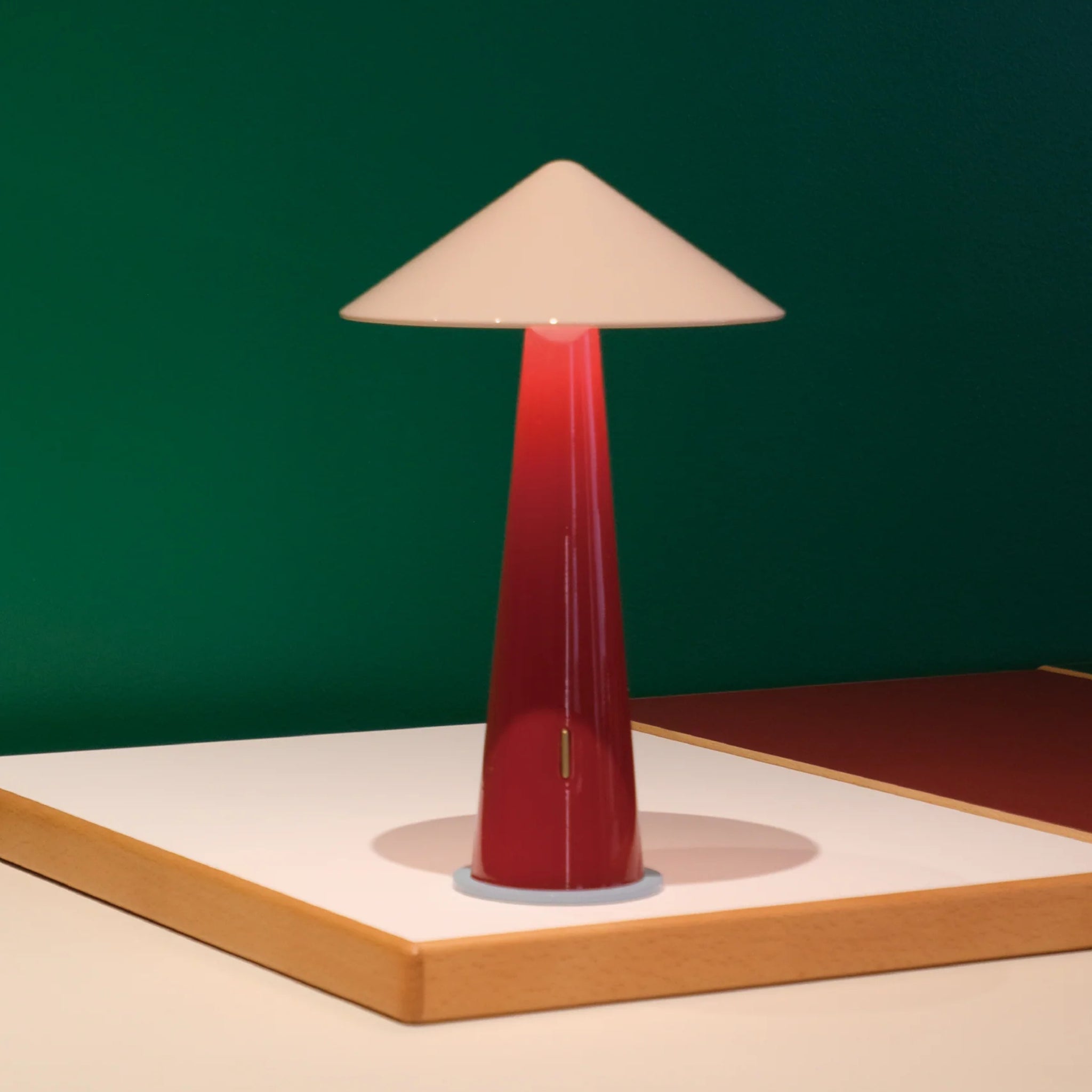 De Fine Lumens Tipsy Lamp LED Oplaadbaar Poederroze Donkerrood, met een hoge glanzende rode voet en kegelvormige crèmekleurige kap, staat op een wit vlak tegen een groene muur.