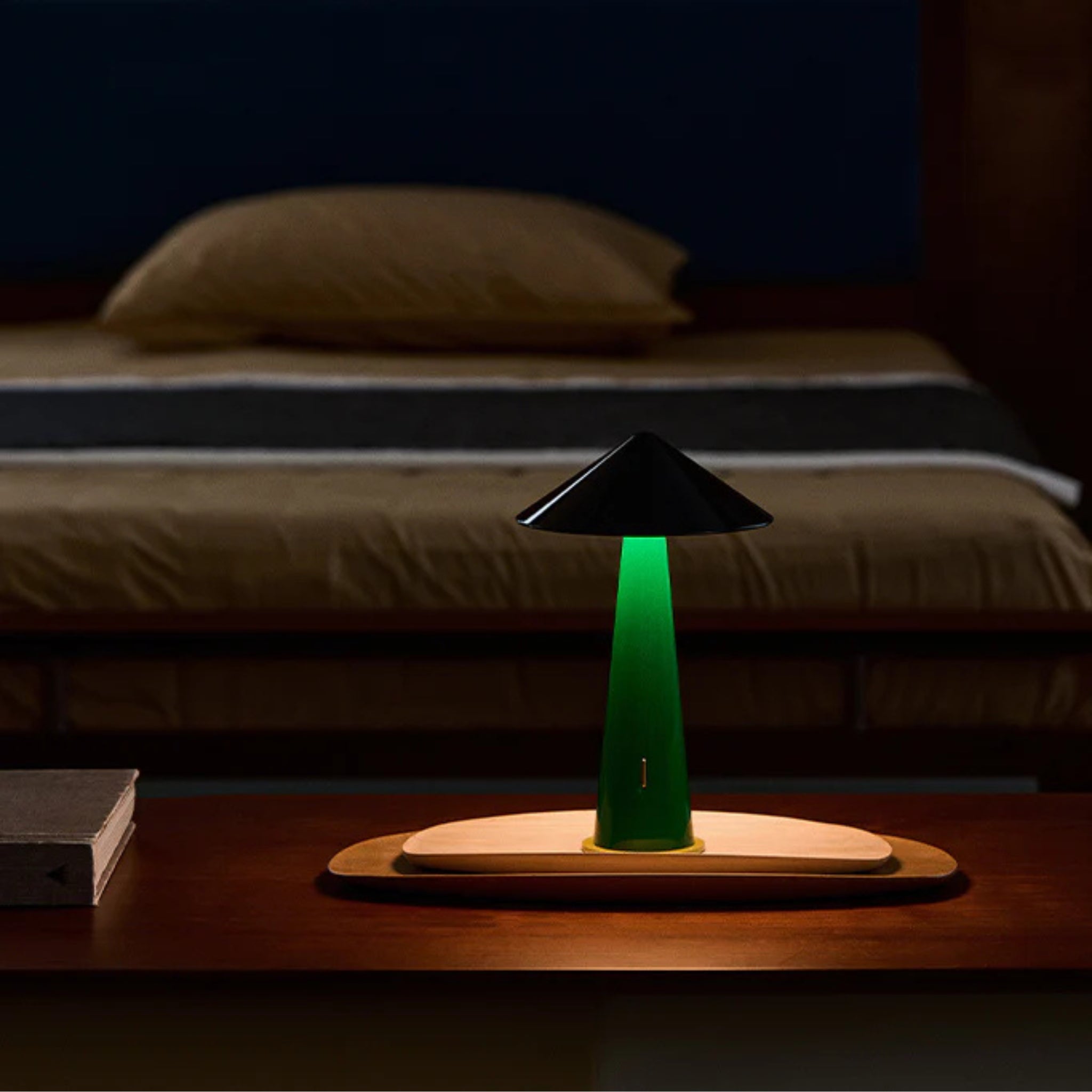 De Fine Lumens Tipsy Lamp LED Oplaadbaar Zwart Emerald Groen gloeit zacht op een houten nachtkastje voor een keurig opgemaakt bed met bruin en grijs beddengoed in een schemerige slaapkamer.