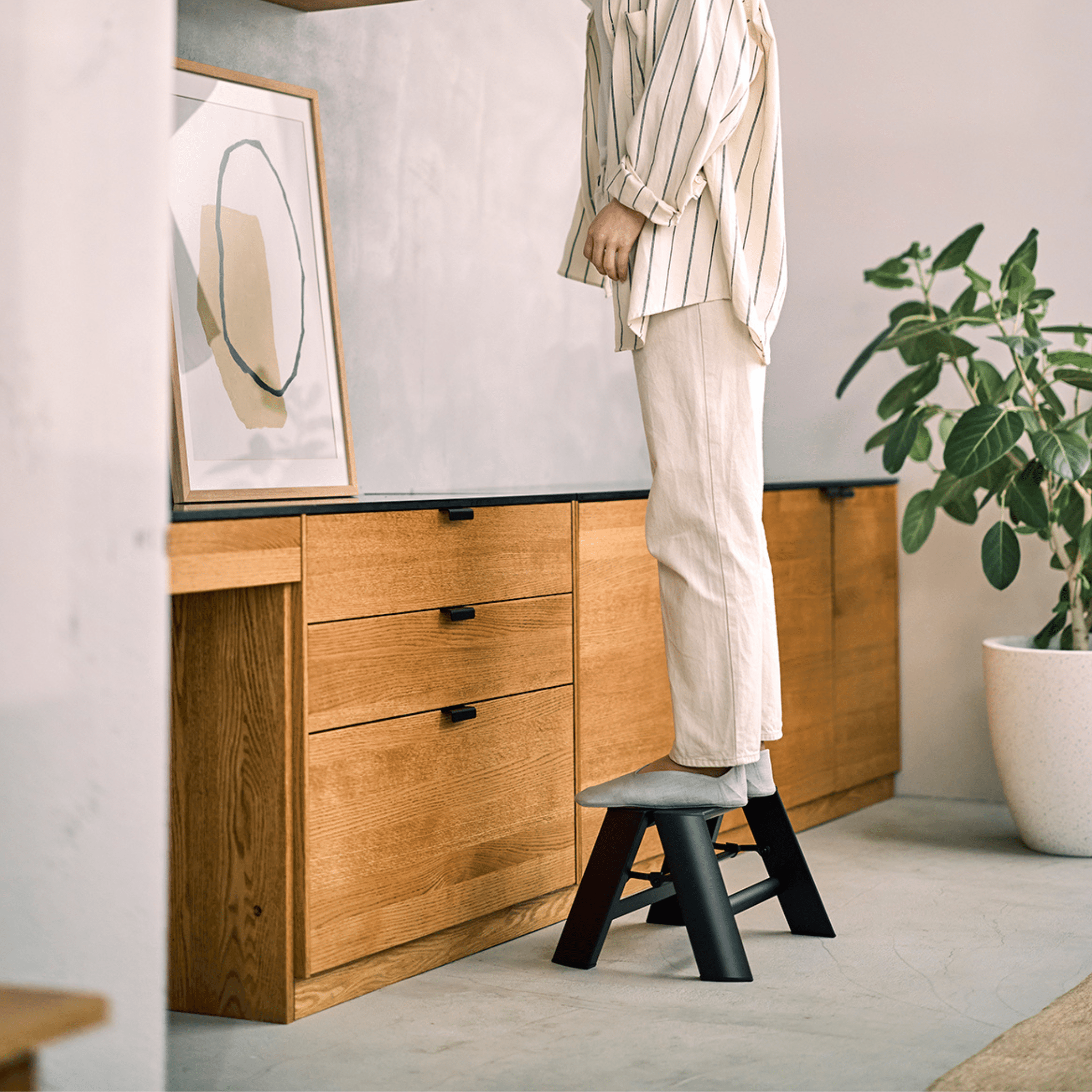 Een persoon in lichte kleding gebruikt de Hasegawa Lucano Ladder 1 Trede Zwart van Hasegawa om bij een houten kast te komen in een moderne, Japans geïnspireerde woning. Een potplant en ingelijste abstracte kunst versieren de ruimte.