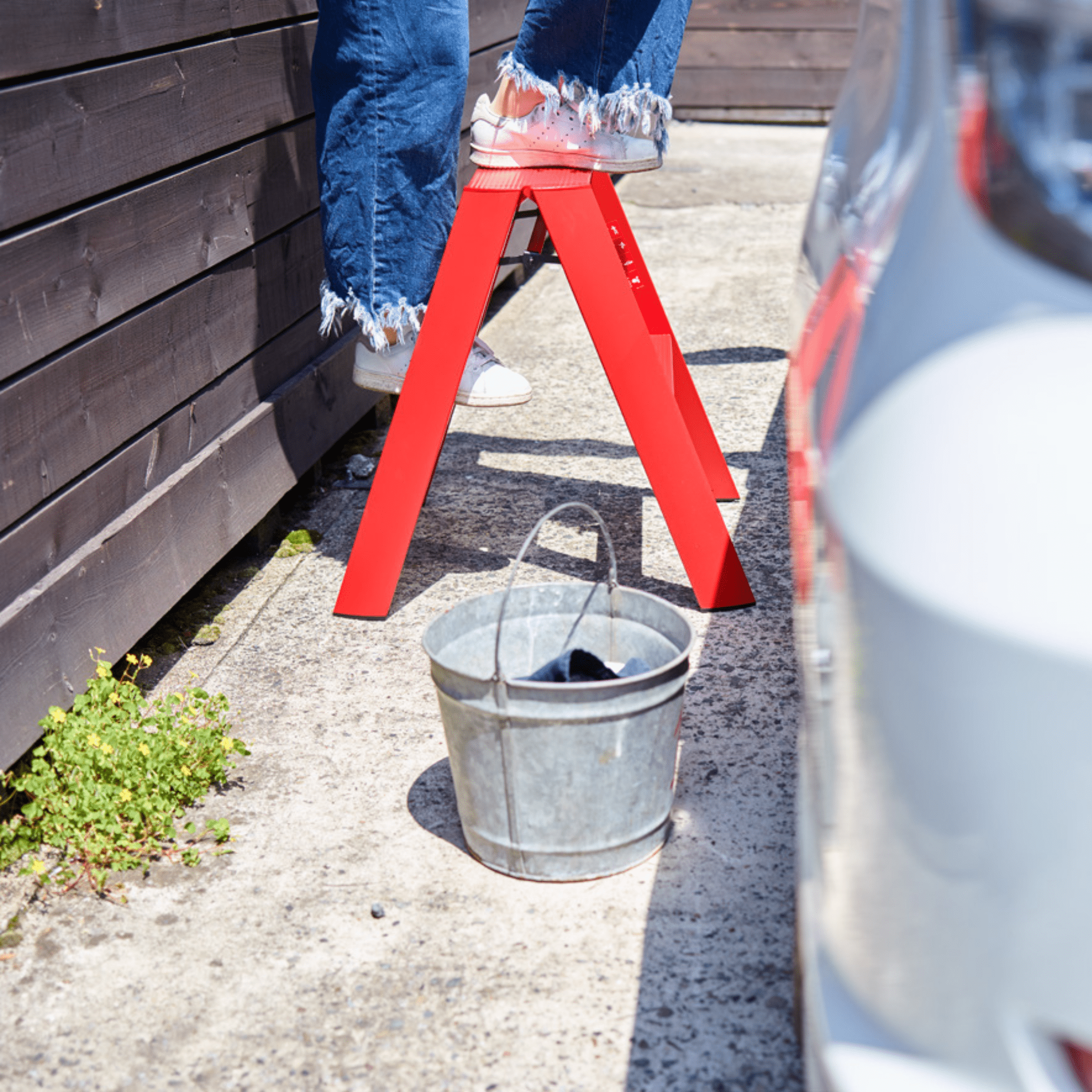 Een persoon in een spijkerbroek met rafels en witte schoenen staat op een Hasegawa Lucano Ladder 2 Step Rood van Hasegawa bij een houten hek, met een metalen emmer op de grond naast een geparkeerde auto.