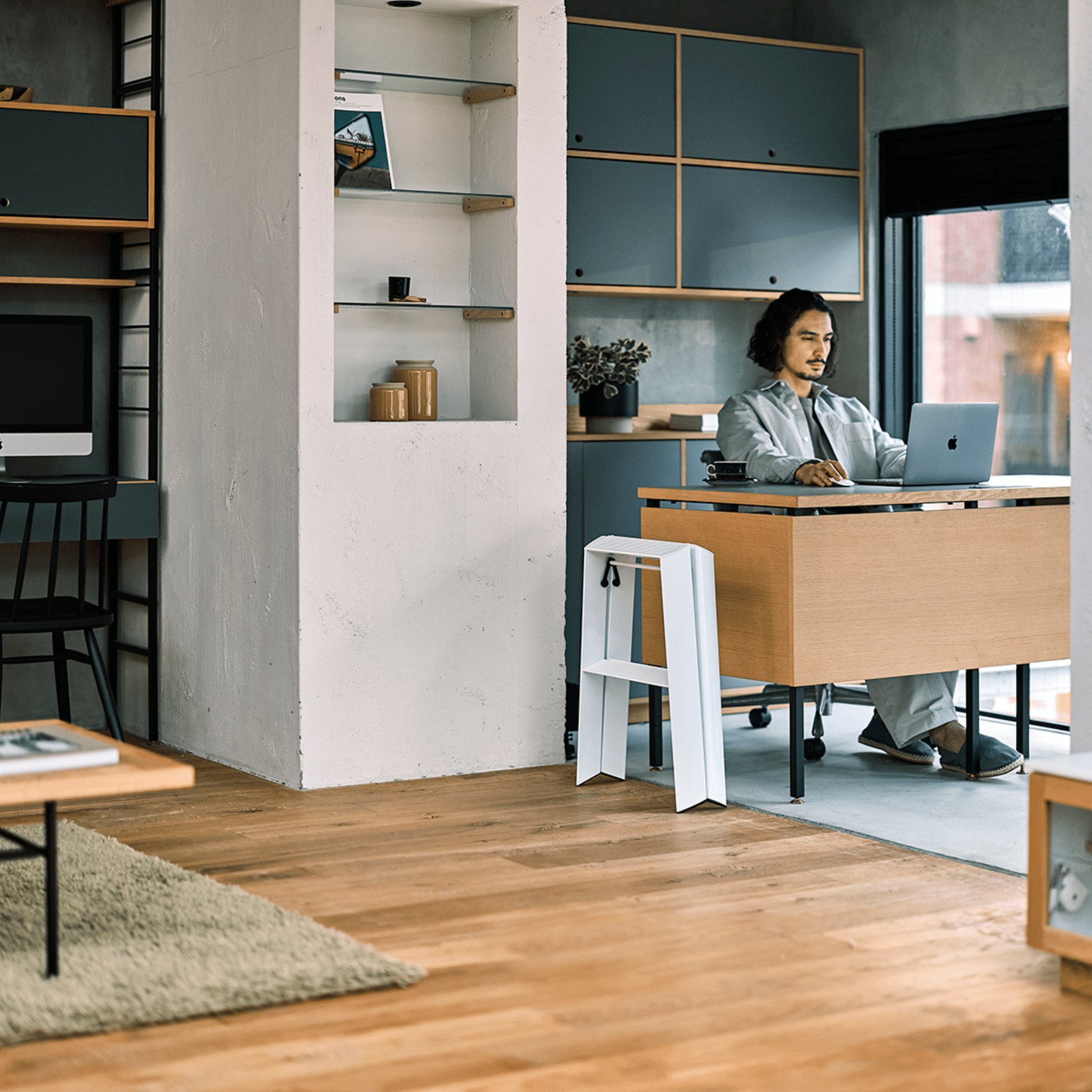 In een minimalistisch kantoor werkt iemand aan een modern bureau terwijl de Hasegawa Lucano Ladder 2 Step Wit van Hasegawa, geïnspireerd op Japans design, vlakbij staat voor gemakkelijke toegang tot hoge planken.