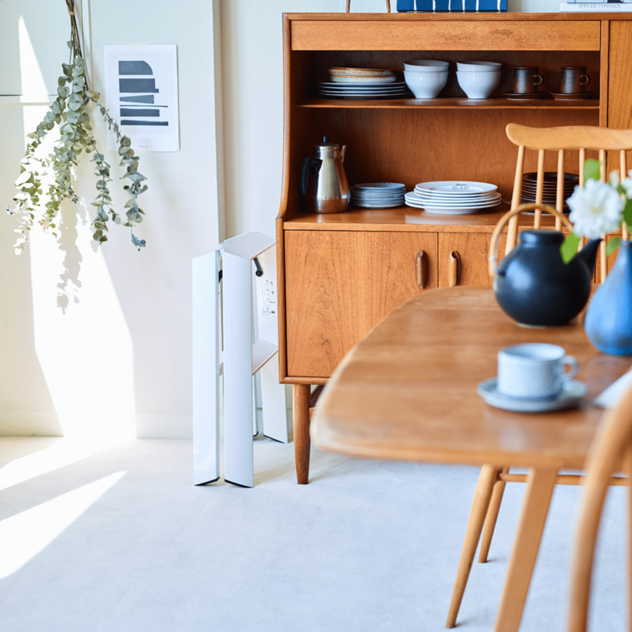 Een lichte eetkamer met Japans design heeft een houten tafel, stoelen, hok met borden en kopjes, zwarte theepot, blauwe kop en schotel, witte luchtreiniger, bladplant, ingelijste kunst, plus een strakke Hasegawa Lucano Ladder 2 Step Wit van Hasegawa.