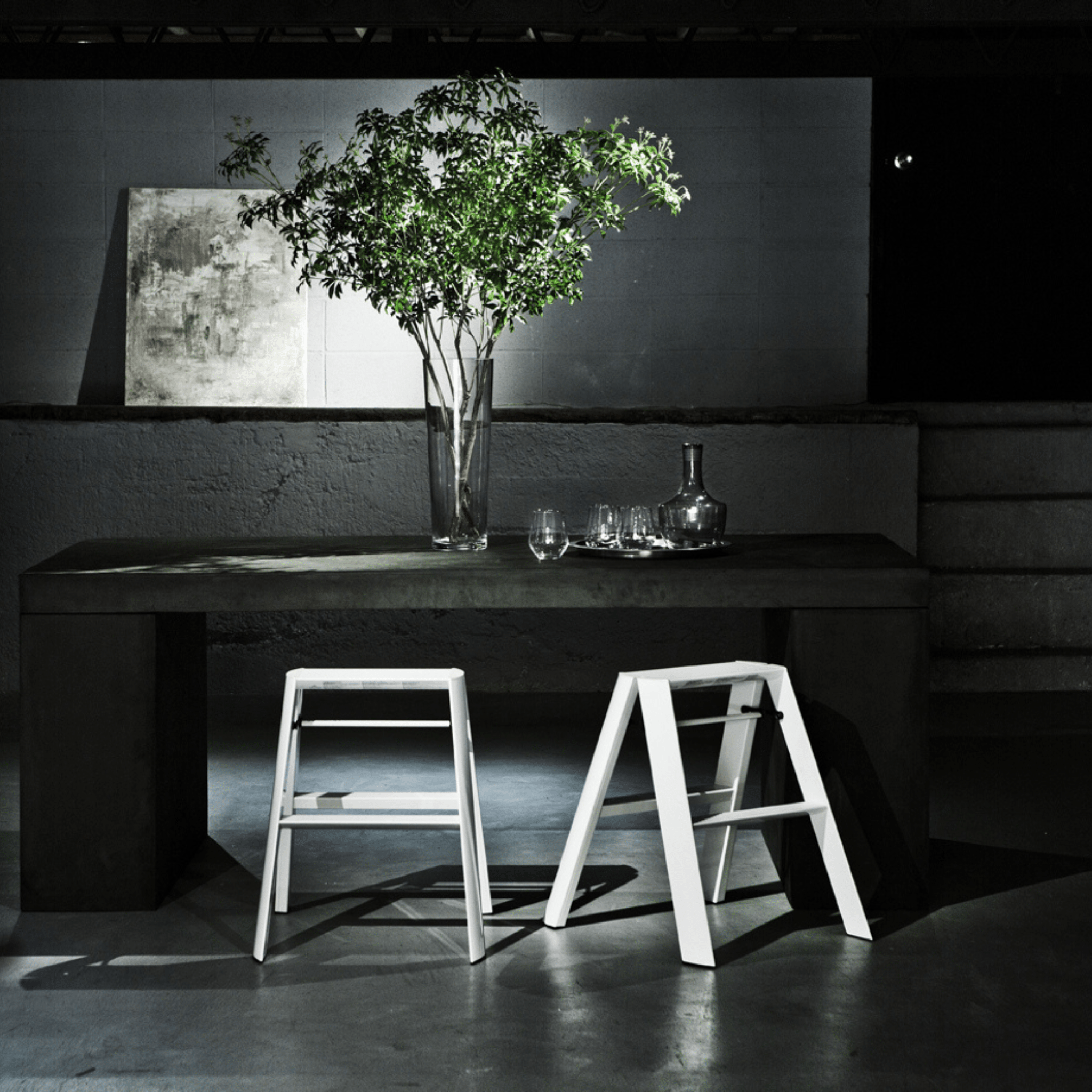 Een minimalistische kamer heeft een donkere rechthoekige tafel met een hoge glazen vaas met groene takken, glaswerk en een karaf. Op de schemerige achtergrond roepen twee Hasegawa Lucano Ladder 2 Step Wit krukken een modern Japans geïnspireerd ontwerp op.