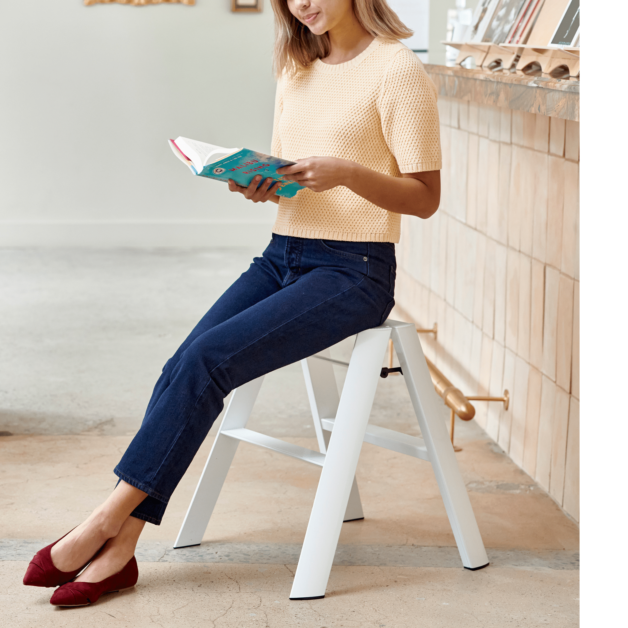 Een vrouw in een lichtgele top en blauwe jeans zit op een Hasegawa Lucano Ladder 2 Step Wit van Hasegawa en leest een turquoise boek. Ze draagt rode platte schoenen binnenshuis bij een toonbank met boeken en decor, die subtiele Japanse designelementen weerspiegelen.