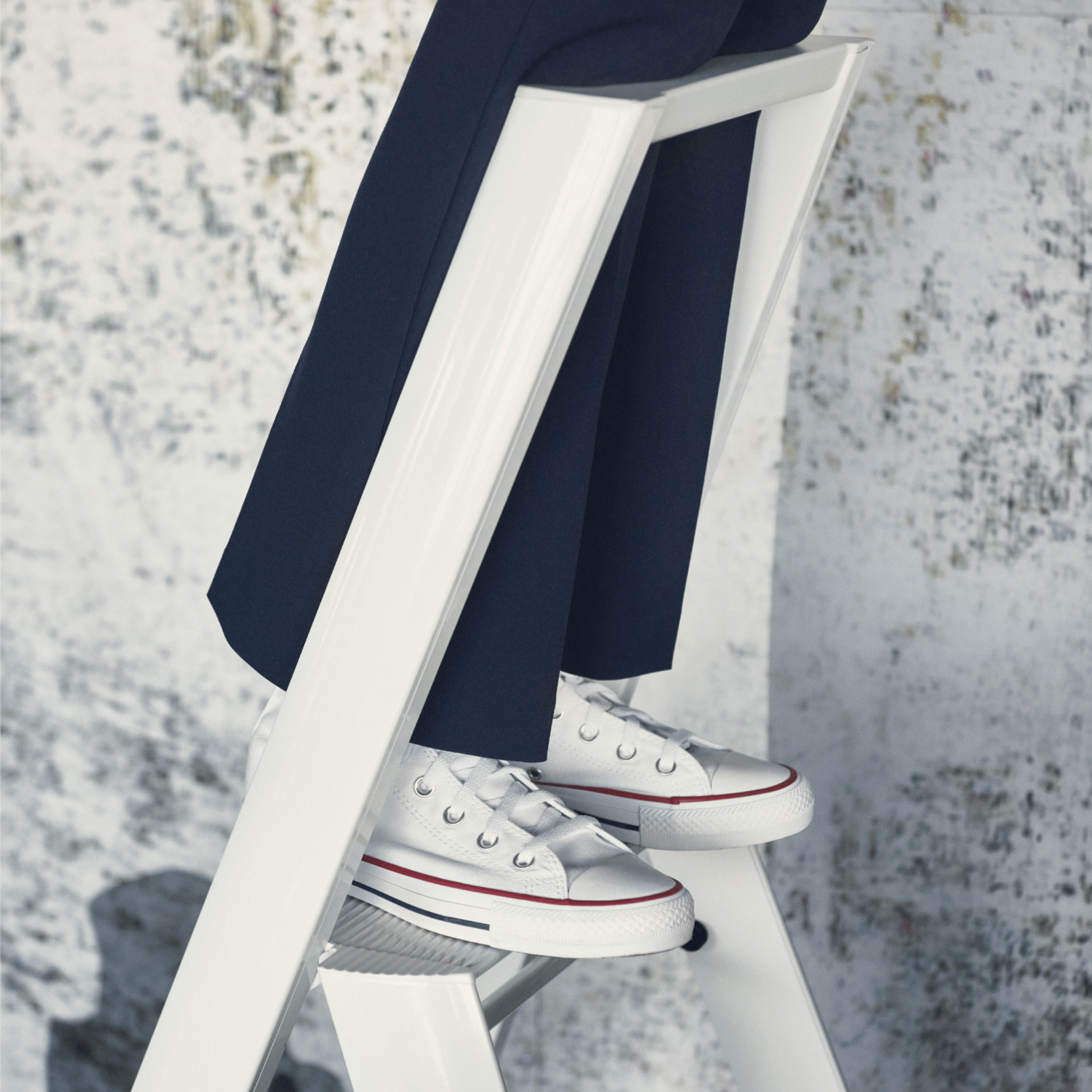 Een persoon in een donkere broek en witte sneakers met rode en blauwe accenten staat op een Hasegawa Lucano Ladder 3 Step Wit van Hasegawa, ontworpen in Japan, tegen een lichtgekleurde muur met textuur.