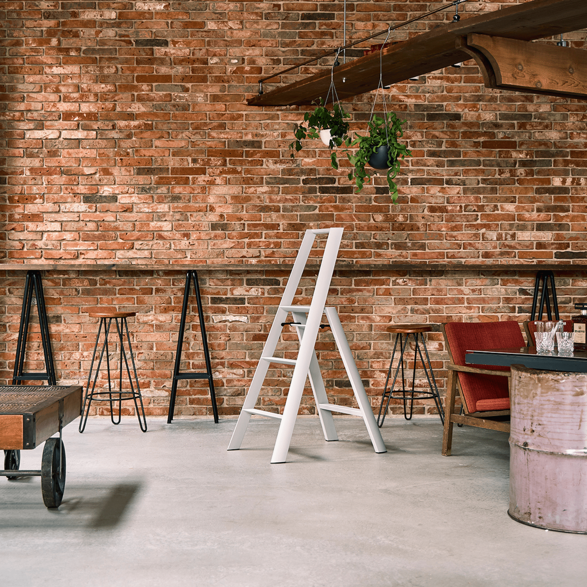 Een modern loftcafé met zichtbare bakstenen muren, hoge krukken, een houten toonbank, de Hasegawa Lucano Ladder 3 Step Wit van Hasegawa in het midden geïnspireerd op Japans design, hangende planten, rode stoelen met kussens en een metalen drumtafel op gepolijst beton.