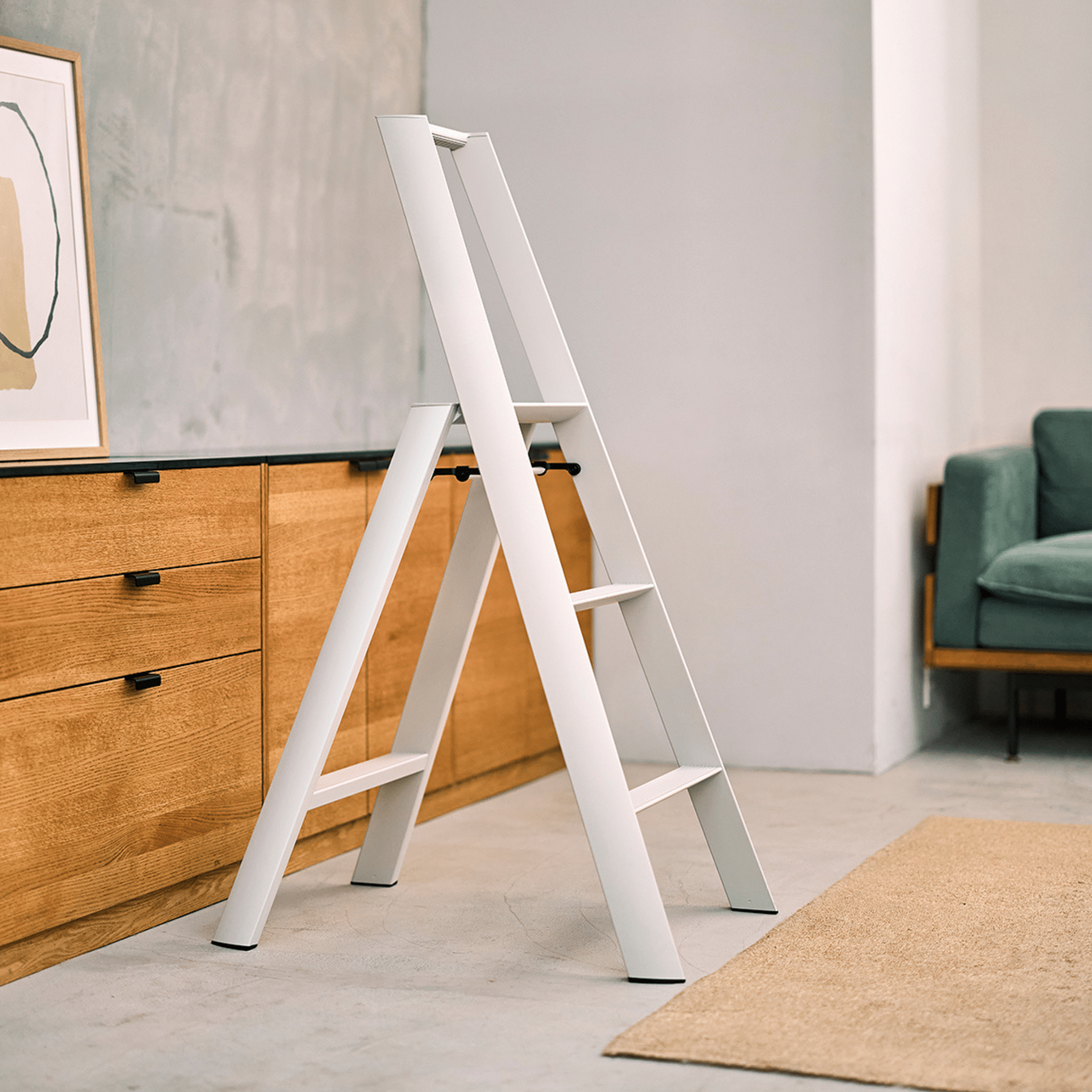 De Hasegawa Lucano Ladder 3 Step Wit van Hasegawa staat open op een lichte vloer naast houten kasten en een groene bank in een minimalistische woonkamer met Japans geïnspireerd design; een abstract kunstwerk leunt tegen de linkermuur.