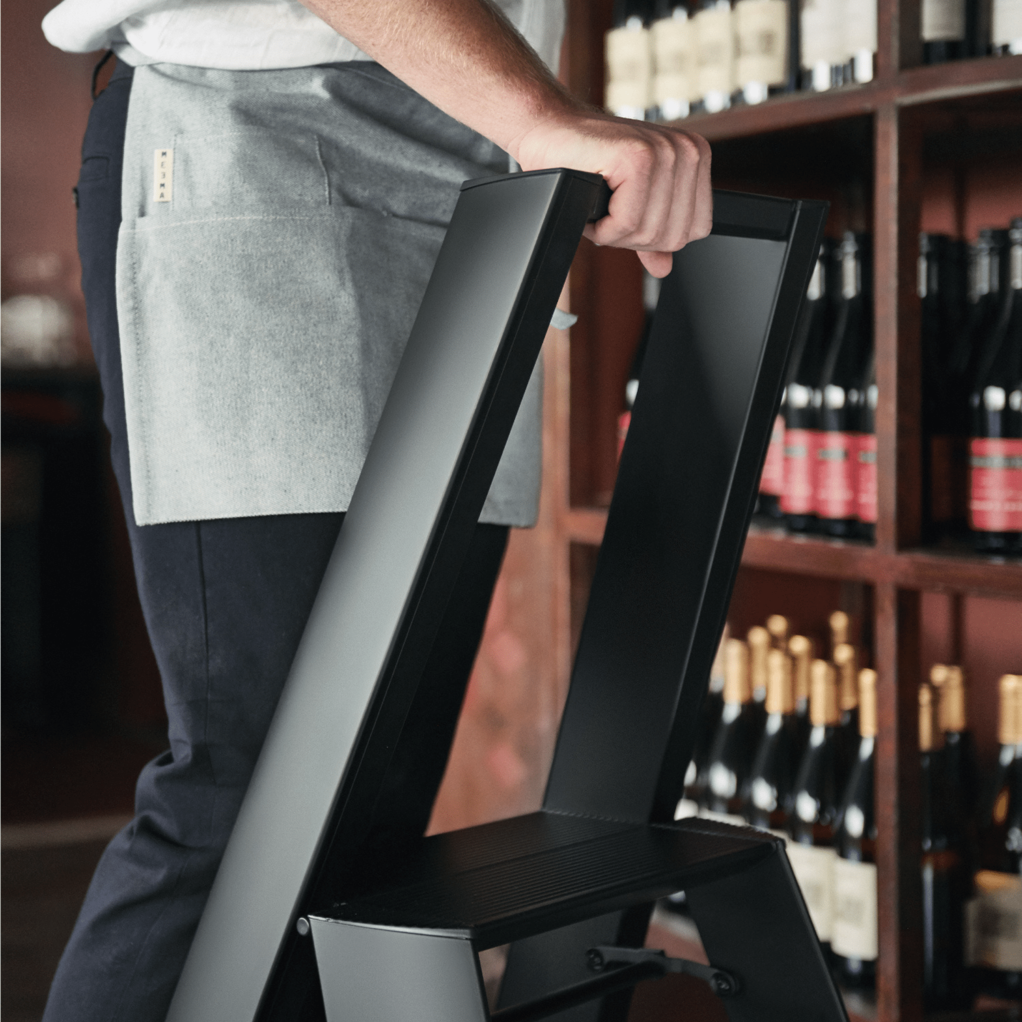 Een persoon in een grijs schort en donkere broek houdt de Hasegawa Lucano Ladder 3 Step Zwart Breed van Hasegawa, een slanke ladder geïnspireerd op Japan, voor planken met wijnflessen.