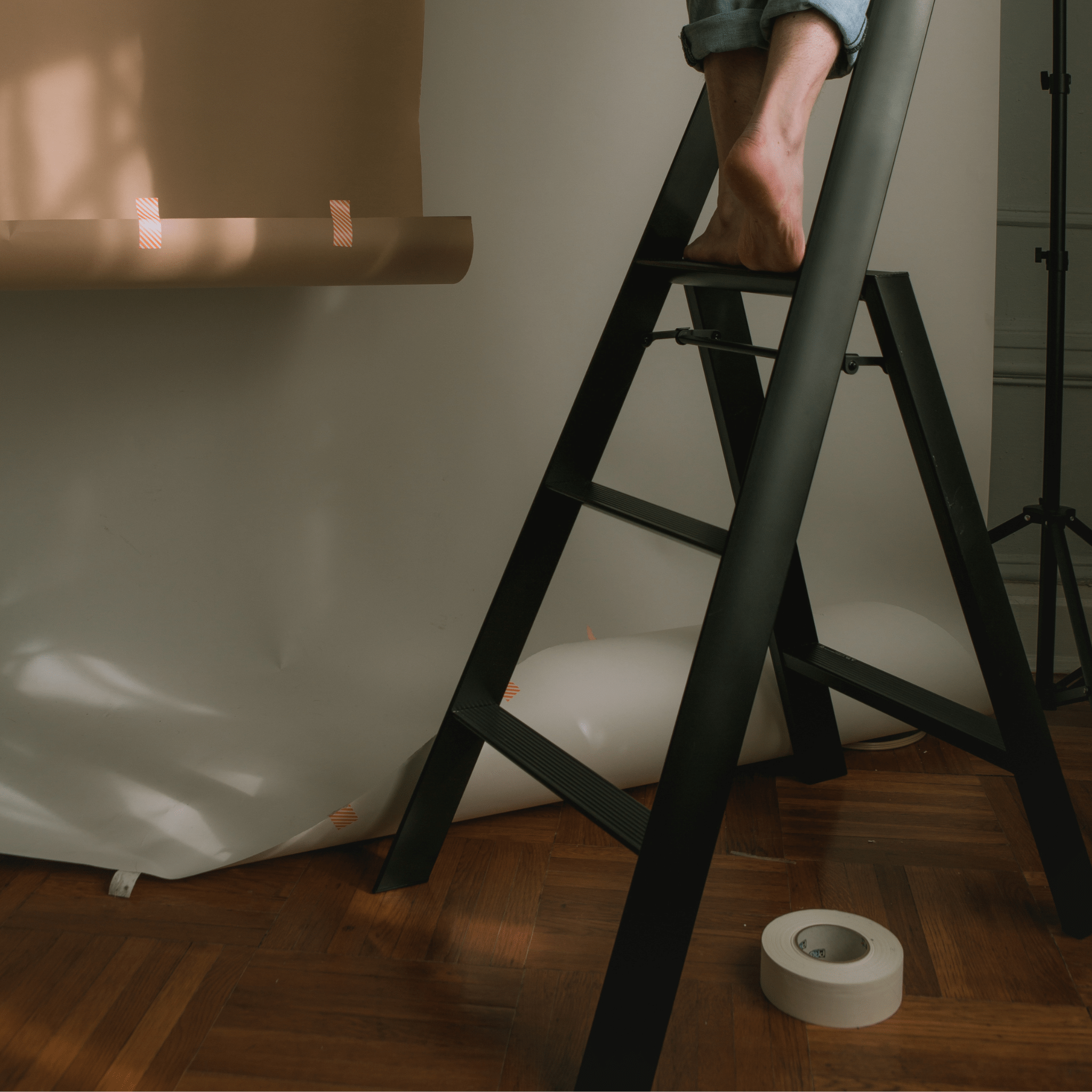 Een persoon met blote voeten staat op een Hasegawa Lucano Ladder 3 Stap Zwart Breed van Hasegawa naast een rol bruin papier die is vastgeplakt aan een witte muur. Een rol plakband rust op de houten vloer met verspreide stukjes papier en plakband in de buurt, wat minimalistisch design oproept.