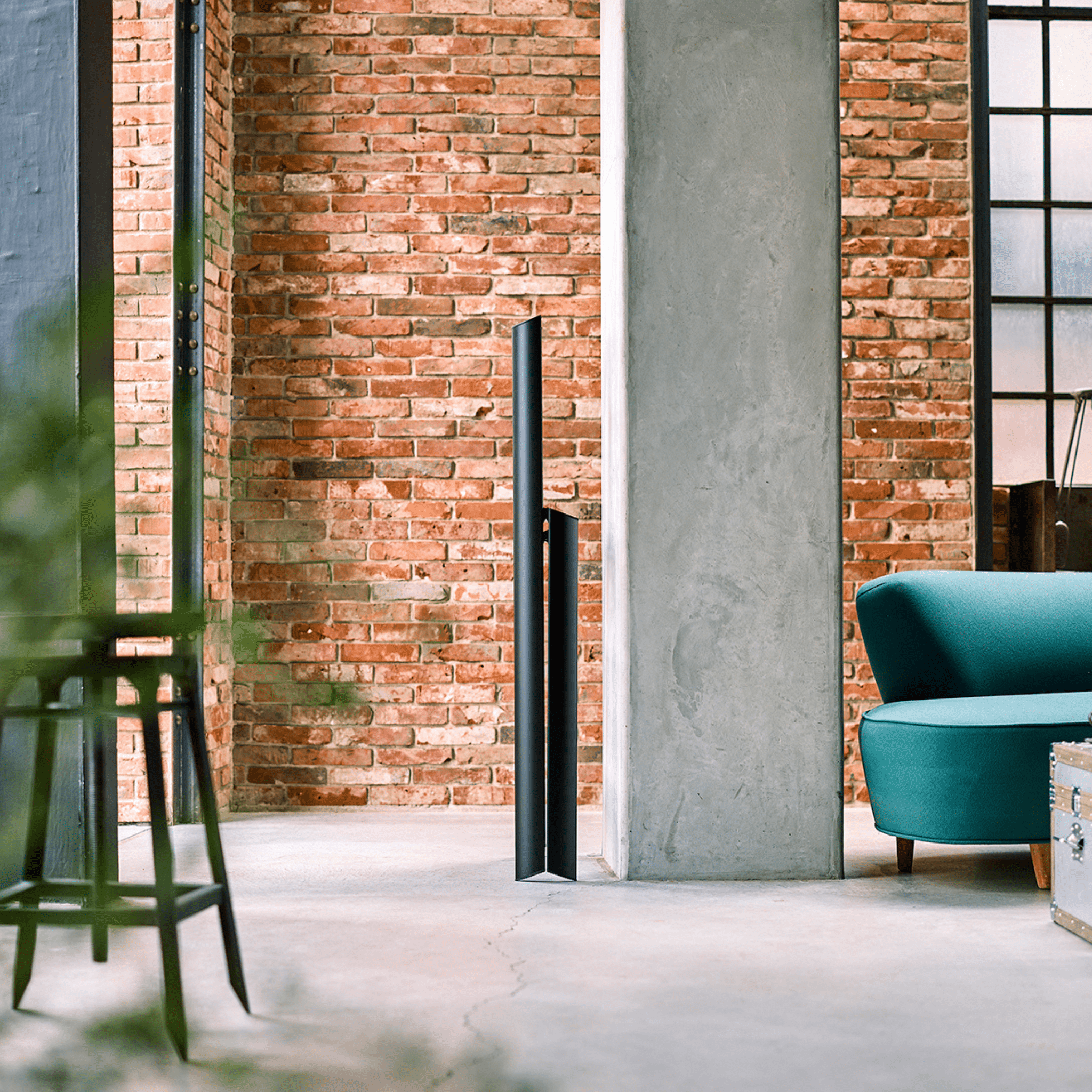 Een minimalistische leefruimte met zichtbare bakstenen, een groenblauwe fauteuil en een vleugje Japans design. Een moderne flatscreentelevisie staat bij de betonnen pilaar, terwijl een Hasegawa Lucano Ladder 3 Step Zwart van Hasegawa gedeeltelijk zichtbaar is aan de linkerkant. Zonlicht valt naar binnen door de grote ramen.