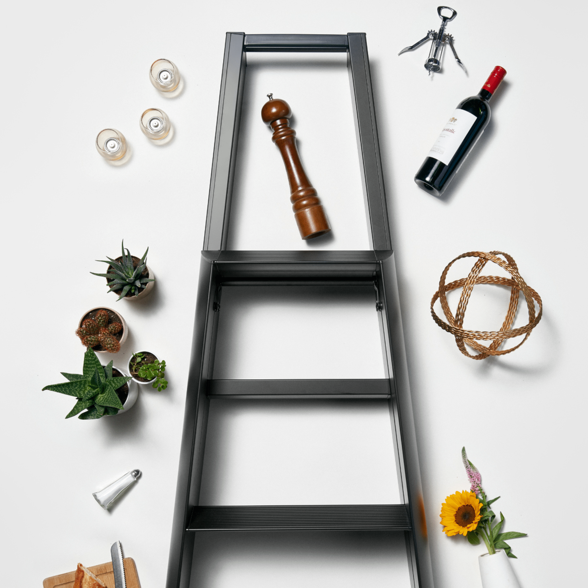 De Hasegawa Lucano Ladder 3 Stap Zwart van Hasegawa staat op een witte achtergrond, omringd door een wijnfles, kurkentrekker, pepermolen, kaarsen, potplanten, touw, zonnebloem en keukengerei.