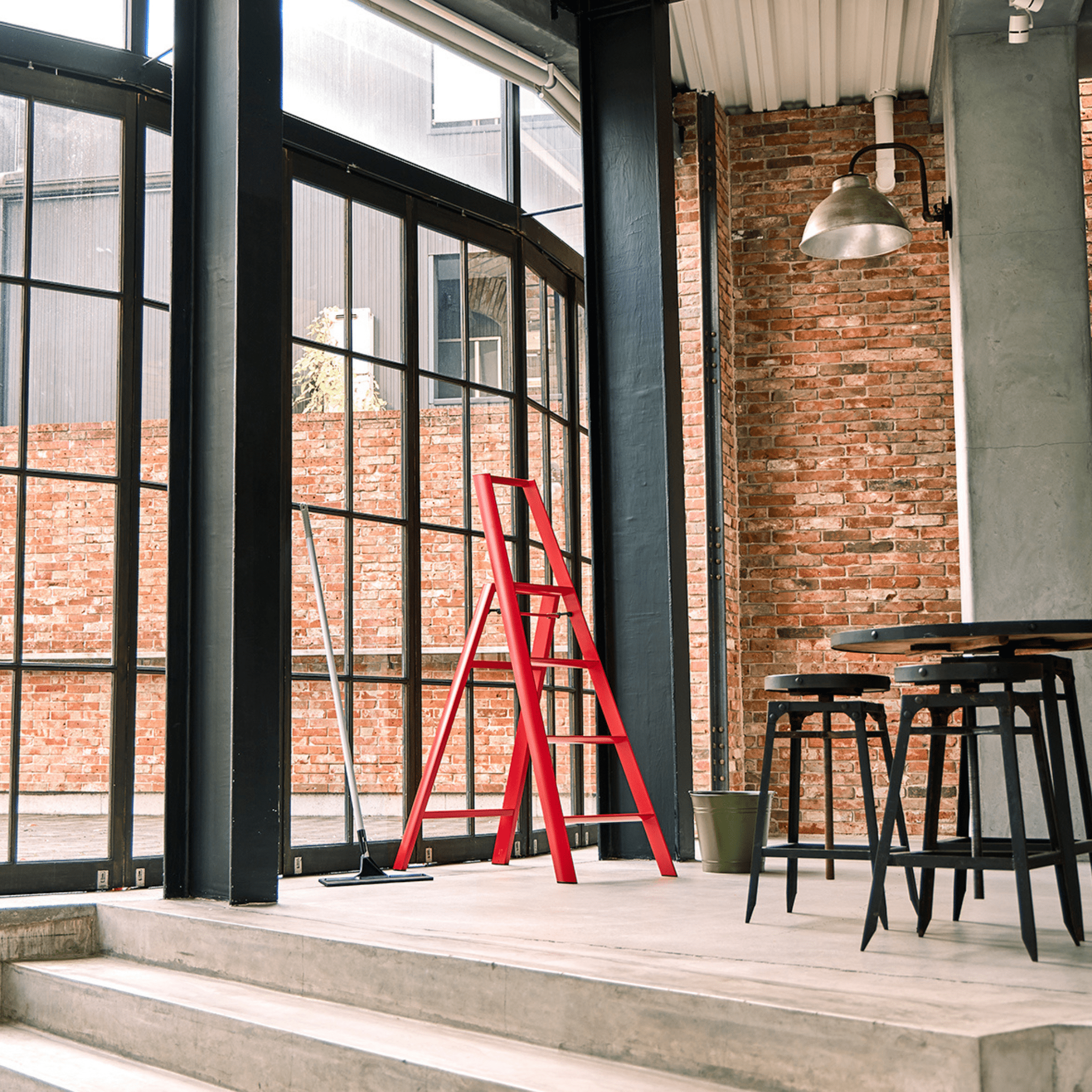 Een moderne kamer in industriële stijl met grote ramen, zichtbare bakstenen, een Hasegawa Lucano Ladder 4 Step Rood van Hasegawa, een dweil, zwarte krukjes rond een hoge tafel en een hangende metalen armatuur met Japans geïnspireerd ontwerp.