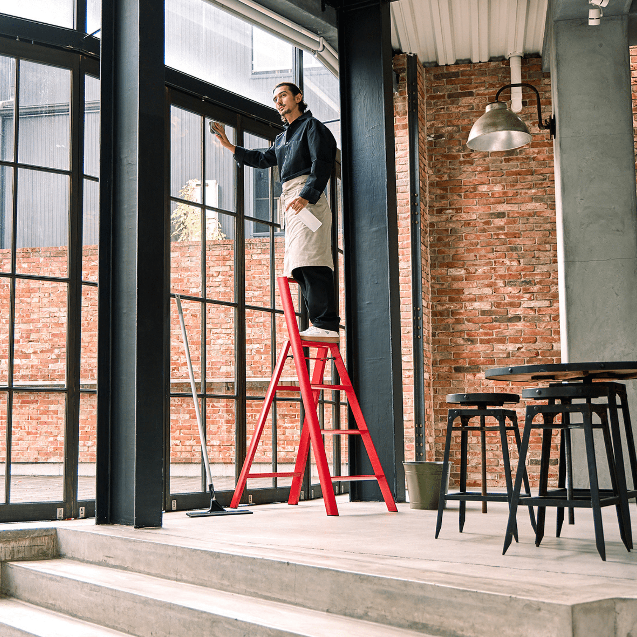 Een persoon gebruikt de Hasegawa Lucano Ladder 4 Stap Rood van Hasegawa om hoge glazen ramen schoon te maken in een moderne, industriële kamer met bakstenen muren, hoge plafonds en Japans geïnspireerd strak zwart meubilair.