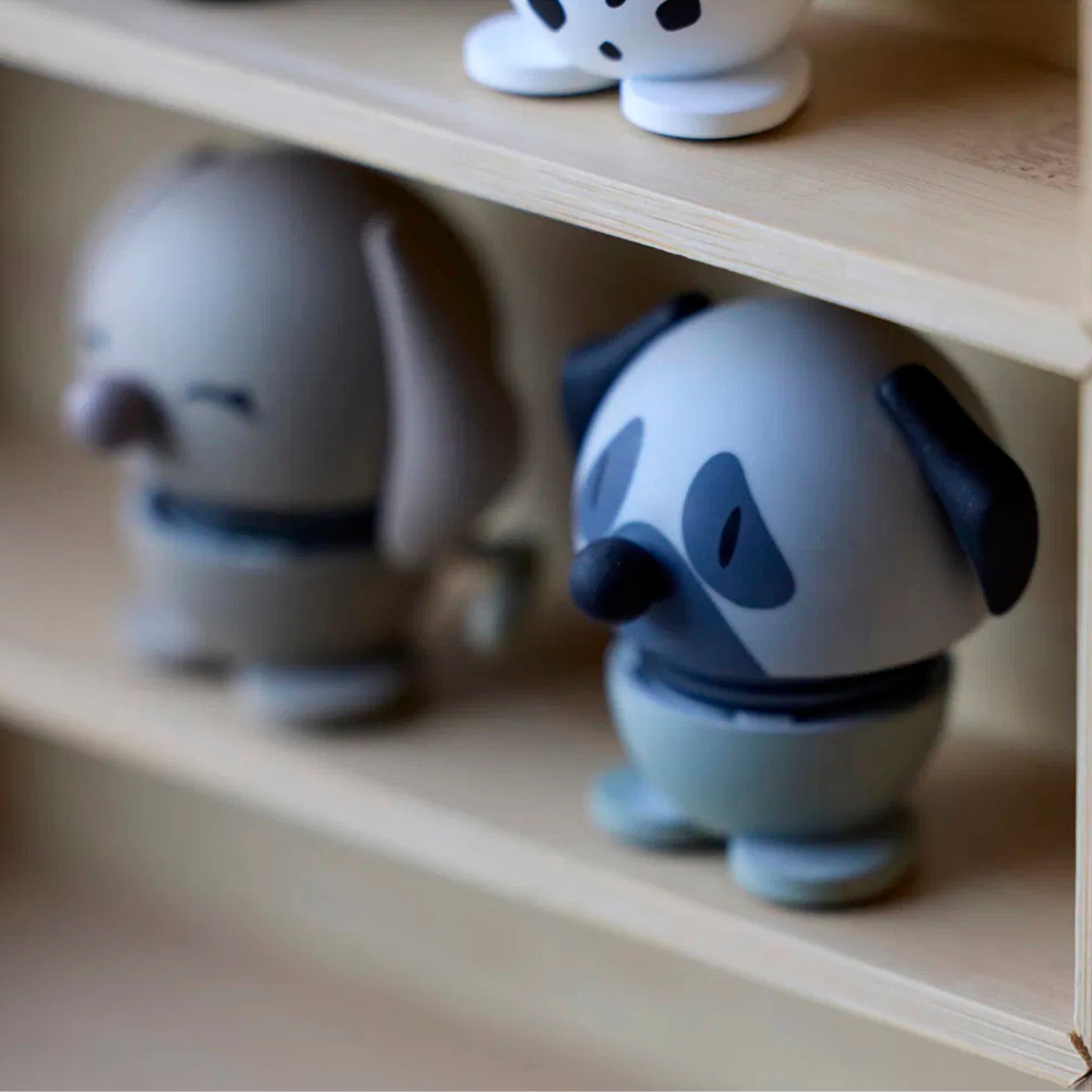 Op een houten plank staan twee kleine, ronde hondenbeeldjes, waaronder de Hoptimist Mopshond Grijs XS van Hoptimist, met beschilderde gezichten en flaporen - een ideaal cadeau voor hondenliefhebbers - met een vage achtergrond en een ander figuurtje erboven.