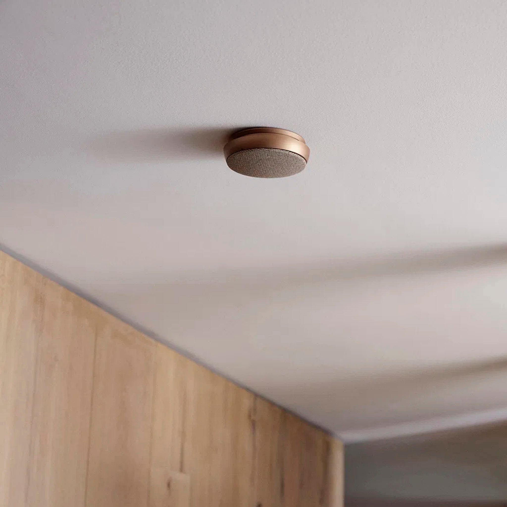 Een Jacob Jensen Optische Rookmelder Koper D10 is gemonteerd aan een wit plafond boven een lichte wand met houten panelen in een moderne, minimalistische kamer.
