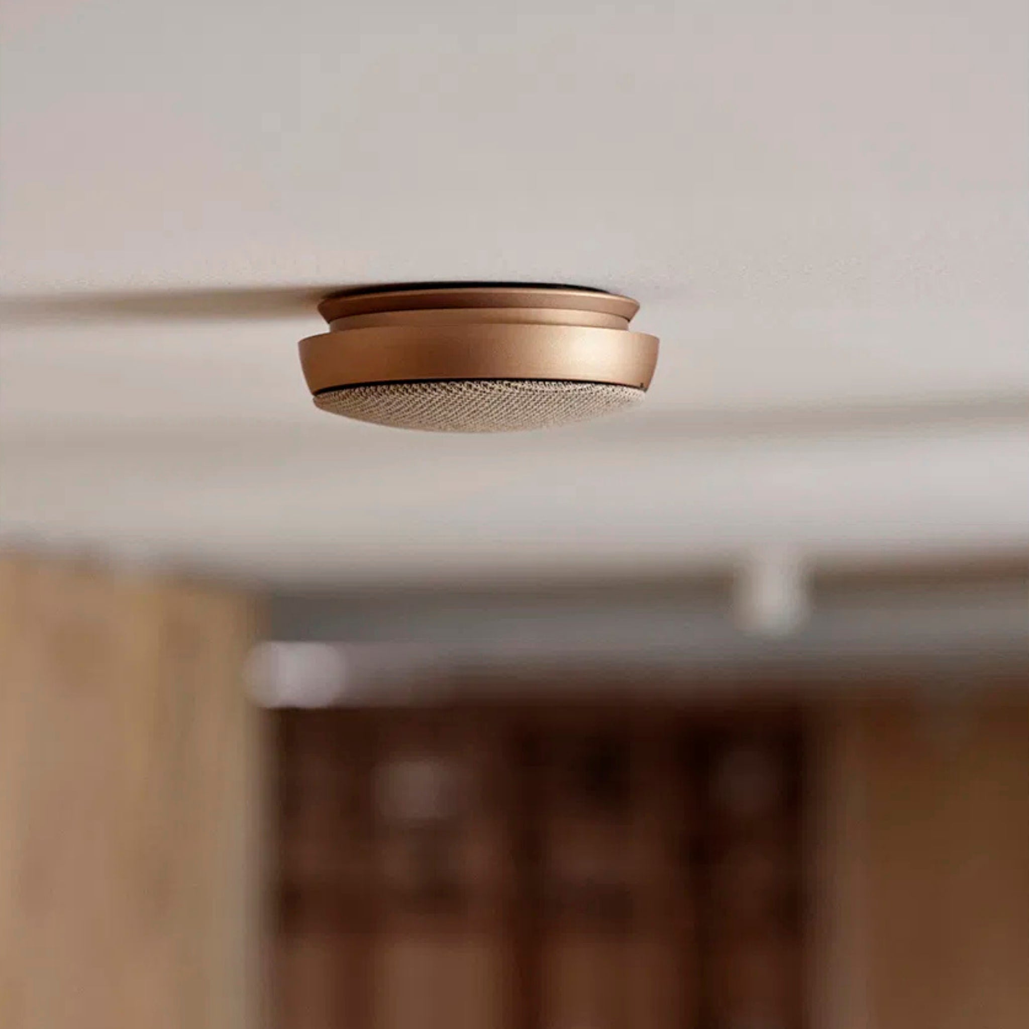 De Jacob Jensen Optische Rookmelder Koper D10 van Jacob Jensen is gemonteerd aan een wit plafond. Het koperkleurige ontwerp steekt elegant af tegen de vage achtergrond van een modern interieur.