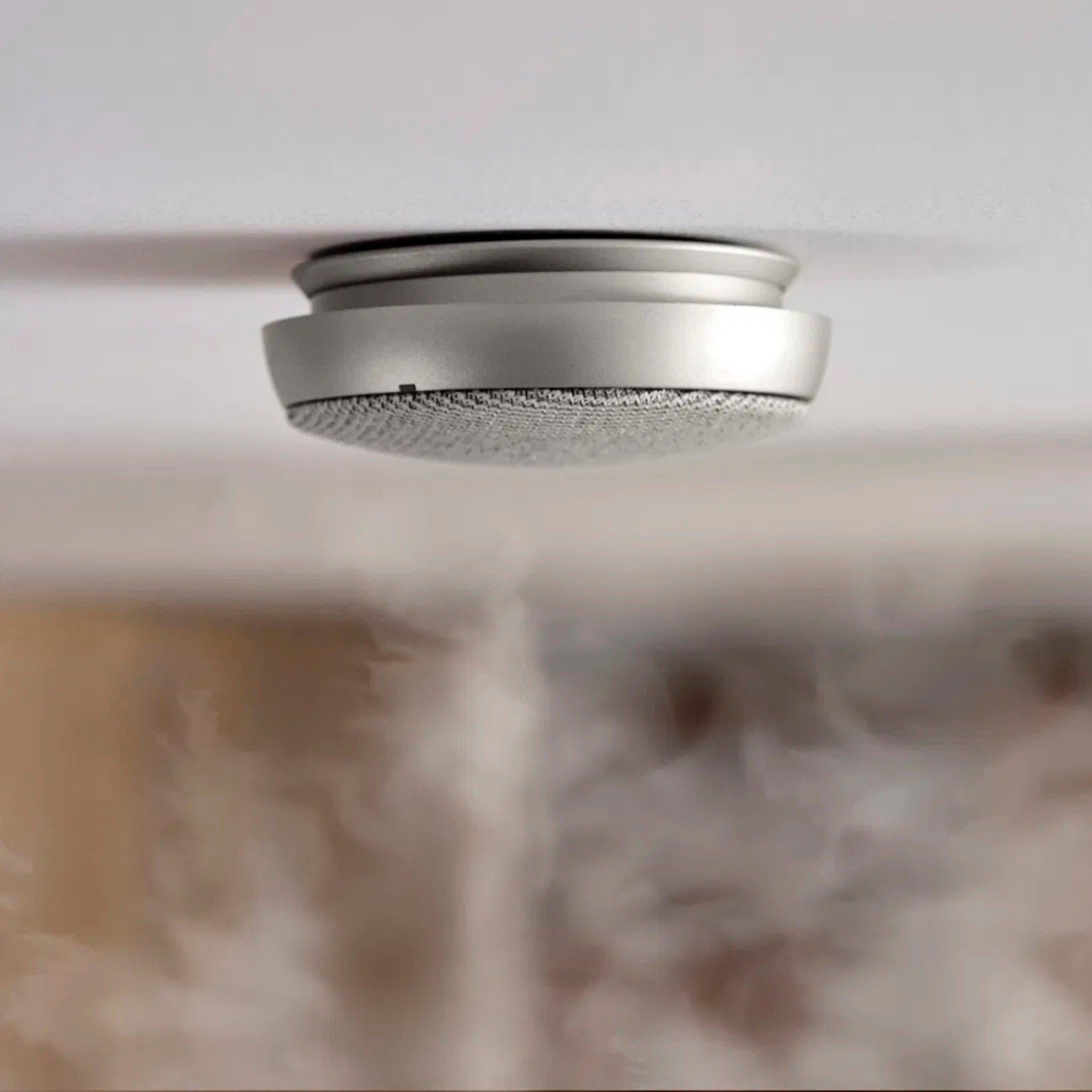 De Jacob Jensen Optical Rookmelder Zilver D10, met Scandinavisch design, is aan een wit plafond gemonteerd terwijl rook opstijgt en deels de achtergrond verbergt.