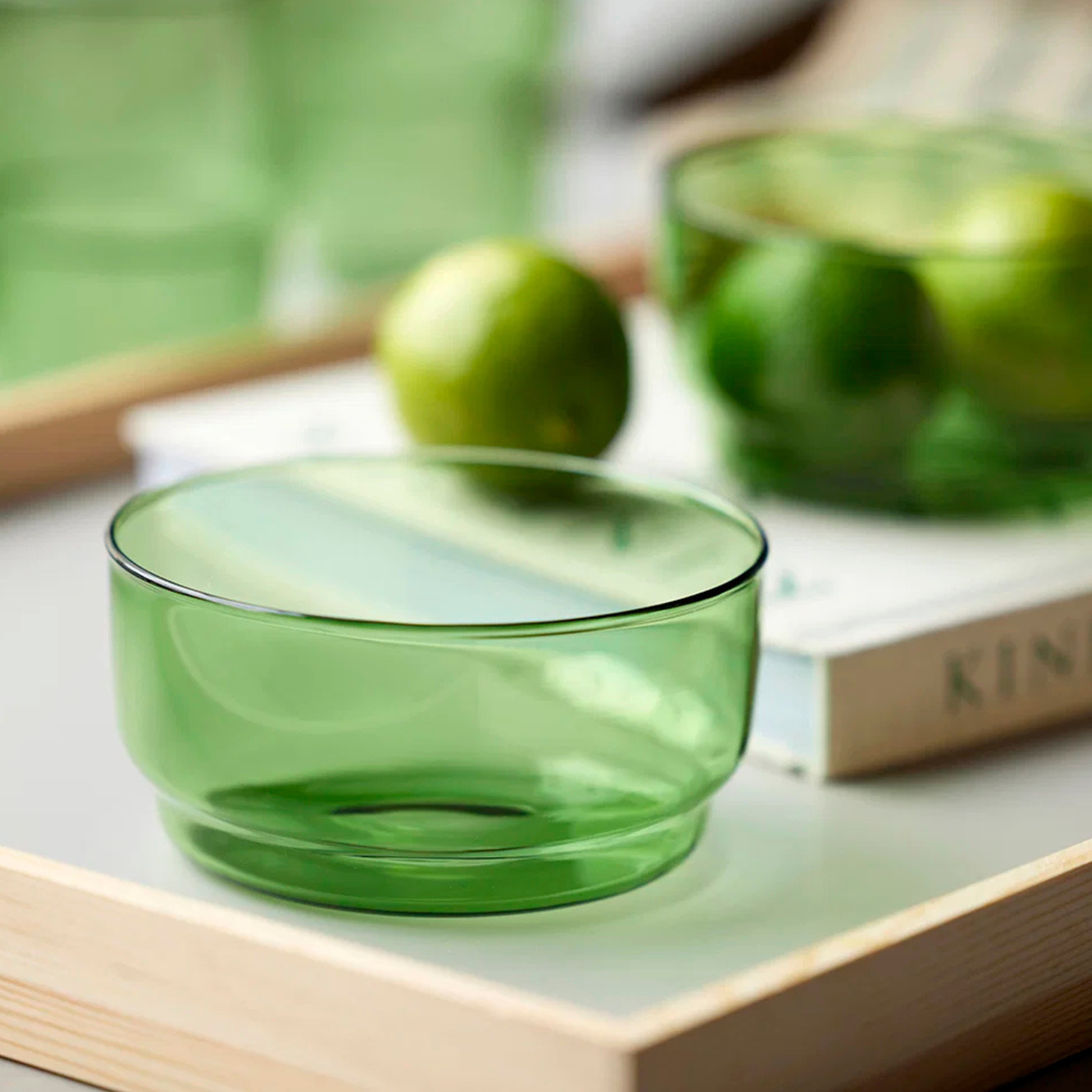 Een Lyngby Glas Torino Kom Groen D12 kom staat op een houten dienblad met een limoen, een andere groene kom gevuld met limoenen en een vaag boek op de achtergrond.