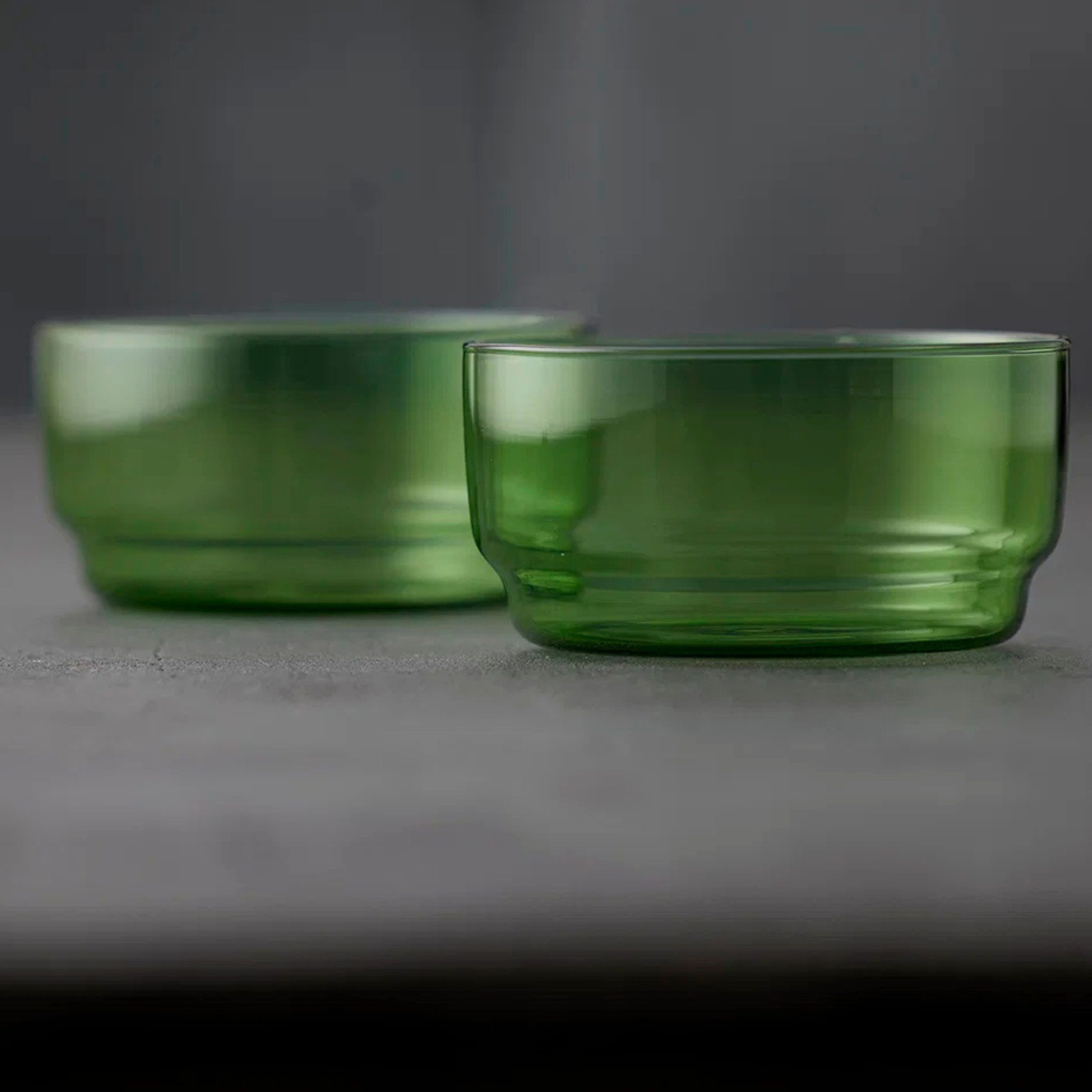 Twee kleine, ronde Lyngby Glas Torino Kom Groen D12 kommen (set van 2) van Lyngby Glas rusten op een grijs oppervlak met een effen donkergrijze achtergrond. De voorste kom is scherp gefocusseerd; de achterste kom is een beetje wazig.