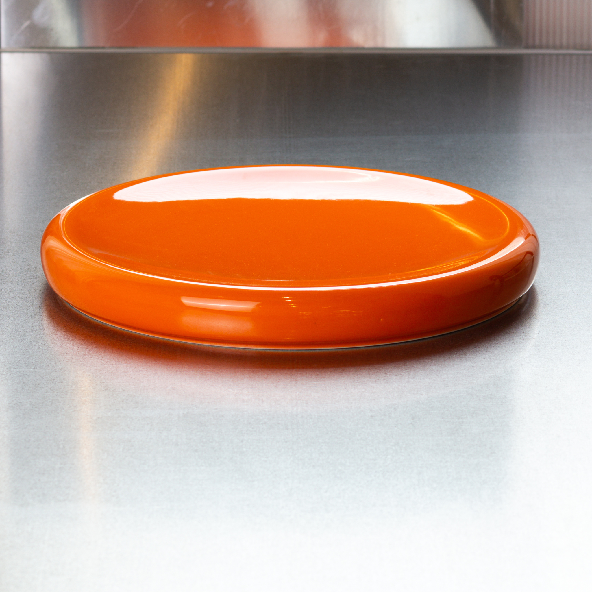 De Matias Moellenbach Big O Serveerschaal Orange, een glanzende, platte porseleinen serveerschaal van Matias Moellenbach, staat op een spiegelend metalen oppervlak met een metalen wand op de achtergrond.