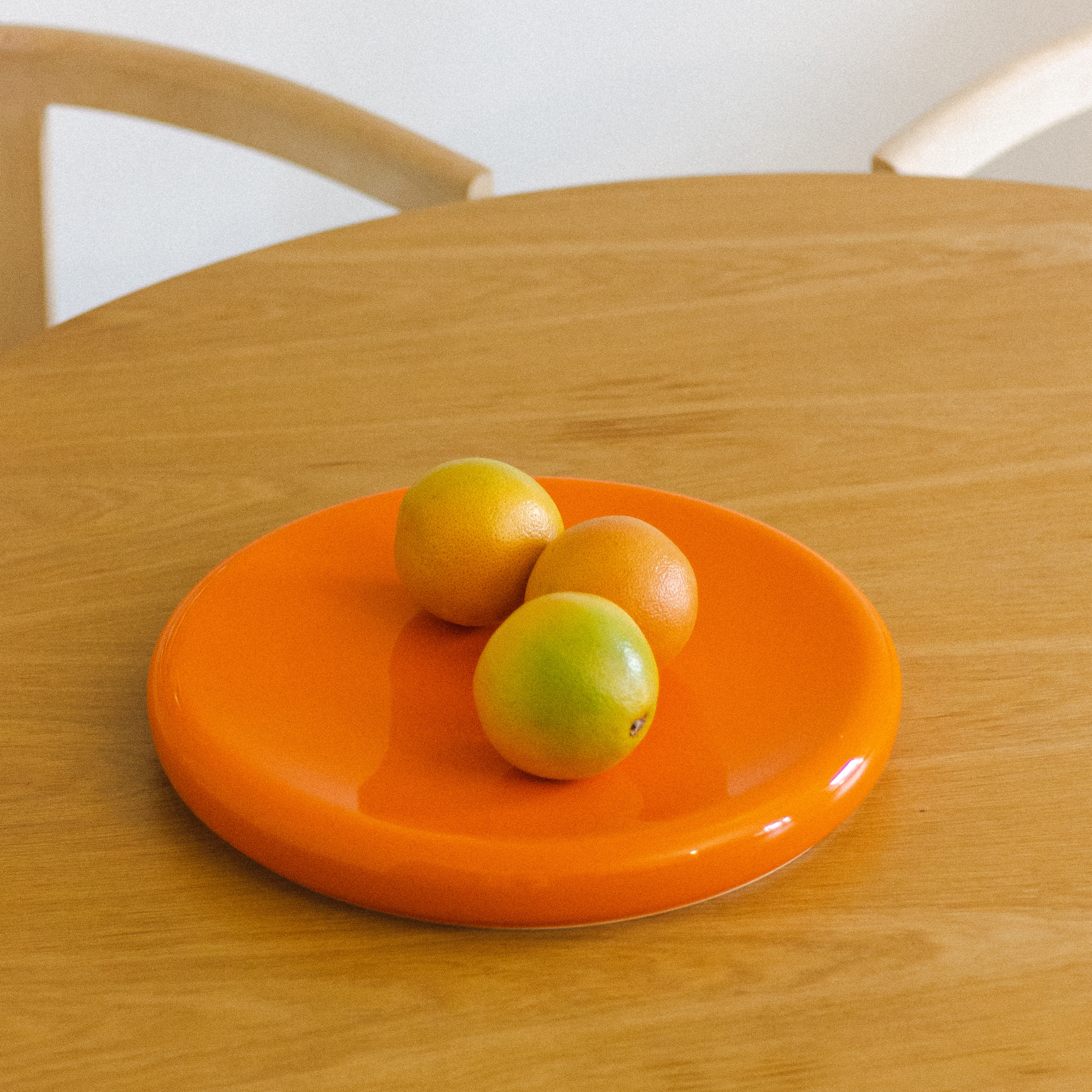 Drie sinaasappels liggen op een „Big O Serveerschaal Orange“ van Matias Moellenbach, die op een lichte houten tafel staat, met op de achtergrond een houten stoel met een gebogen rugleuning.