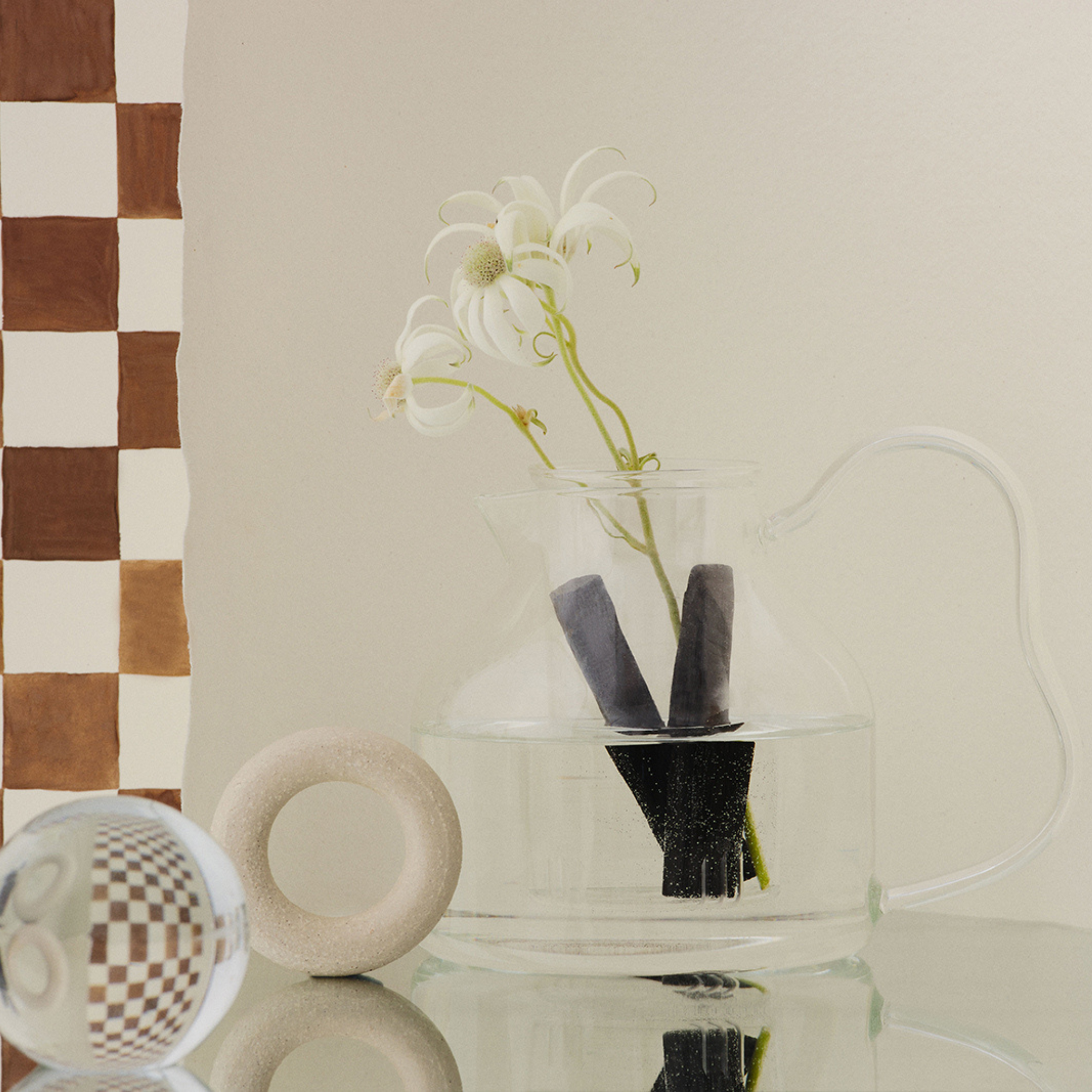 Een heldere glazen kan, versierd met witte bloemen en zwarte stelen, staat op een glazen oppervlak en roept de minimalistische stijl op die kenmerkend is voor Matias Moellenbach. Vlak daarnaast vormen een ronde steen en een geruite bal een aanvulling op het bruin-wit geruite patroon op de muur. Het tafereel straalt zowel elegantie als speelsheid uit en doet denken aan het kunstenaarschap dat tot uiting komt in de Matias Moellenbach Bincho-theepot.