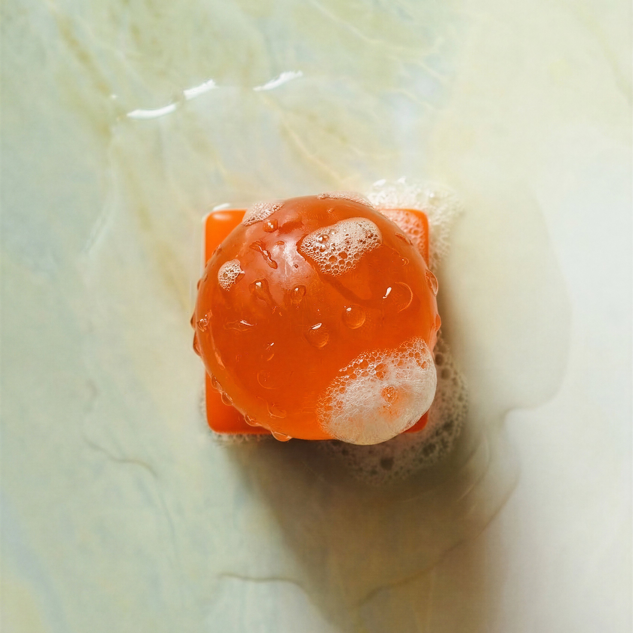 Een oranje bol, die doet denken aan een Matias Moellenbach OBA Zeep Bar Papaya, is bedekt met zeepbellen en staat op een vierkante oranje voet, net als een navulzeep OBA Zeep Houder. Rondom liggen water en zeepsop op een bleek, glad oppervlak.