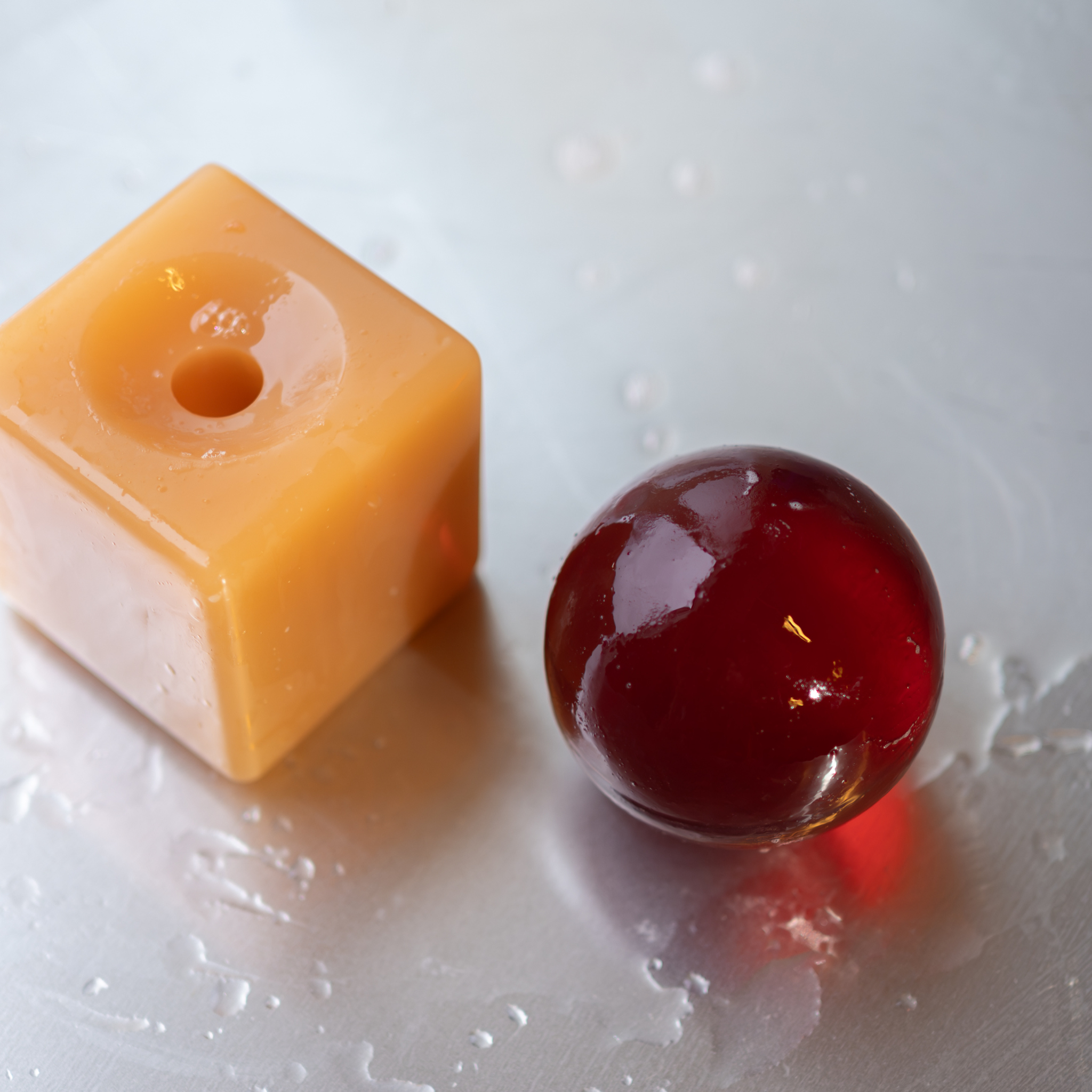 Een gele kubus met een gat en een glanzende rode bol rusten op een nat, metaalachtig oppervlak bezaaid met waterdruppels, waarmee ze de strakke lijnen van het Scandinavische ontwerp weerspiegelen – een esthetiek die terug te vinden is in de Matias Moellenbach OBA Zeep Bar Terralis van Matias Moellenbach.