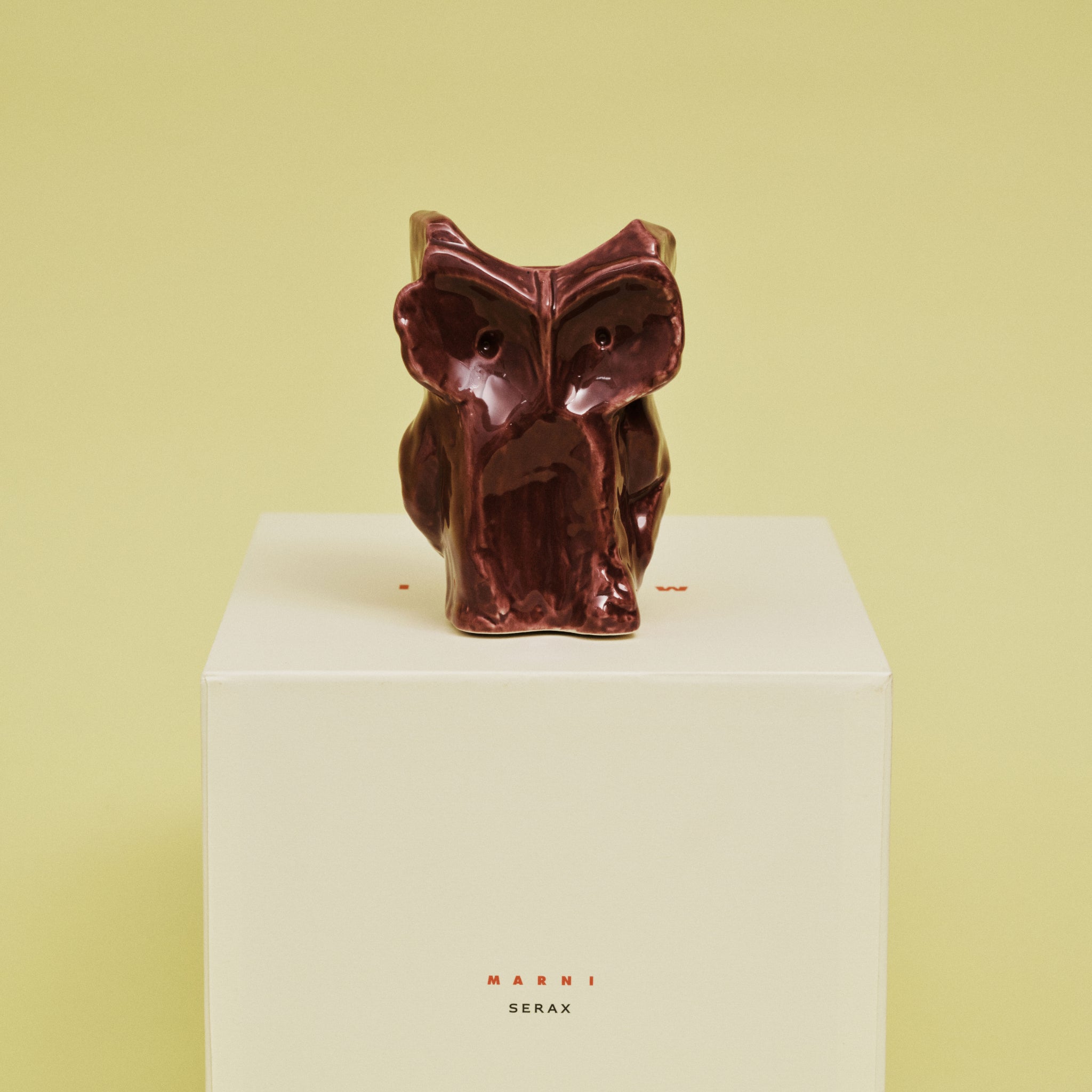 Een glanzende, abstracte Serax After Midnight Uil Vaas Aubergine XS staat op een witte sokkel met "Serax" in rood gedrukt tegen een lichtgele achtergrond.