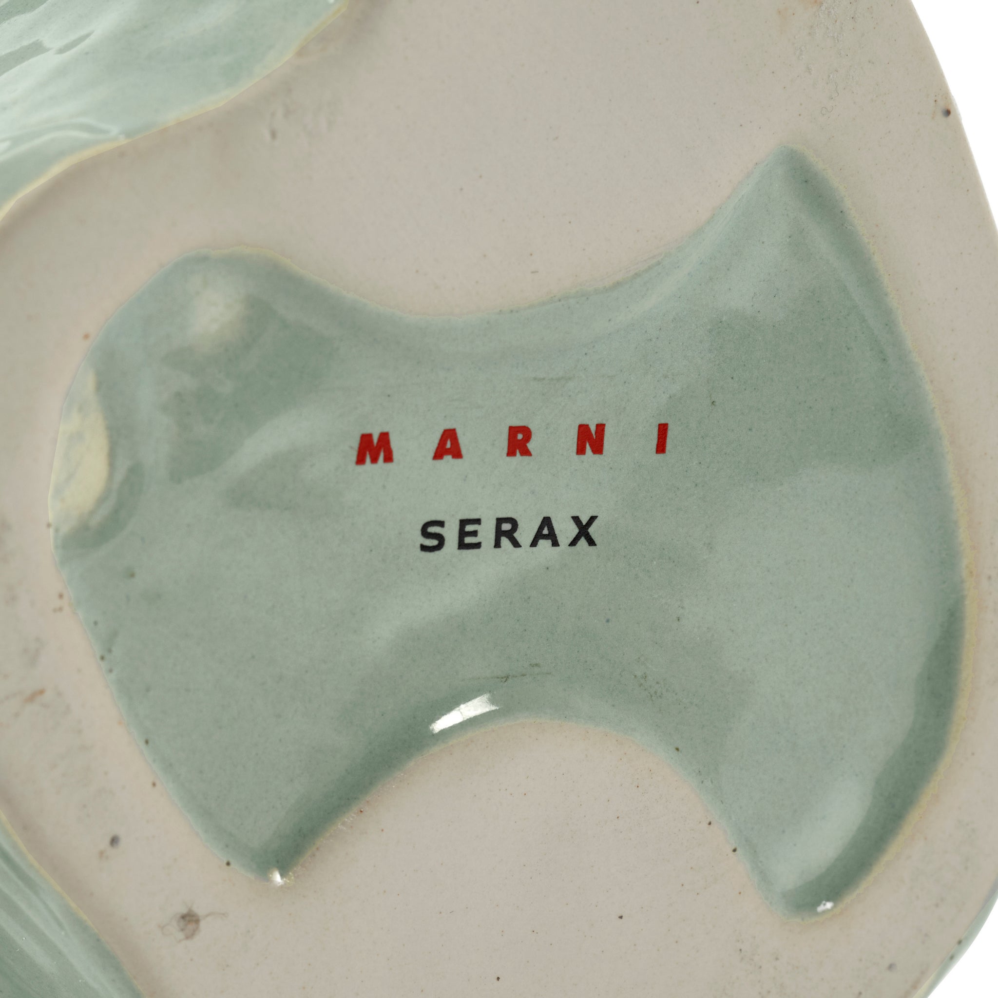 De onderkant van de Serax After Midnight Uil Vaas Lichtblauw XS heeft een glanzende lichtblauwe en beige afwerking, met de merknaam SERAX gedrukt in zwarte hoofdletters.