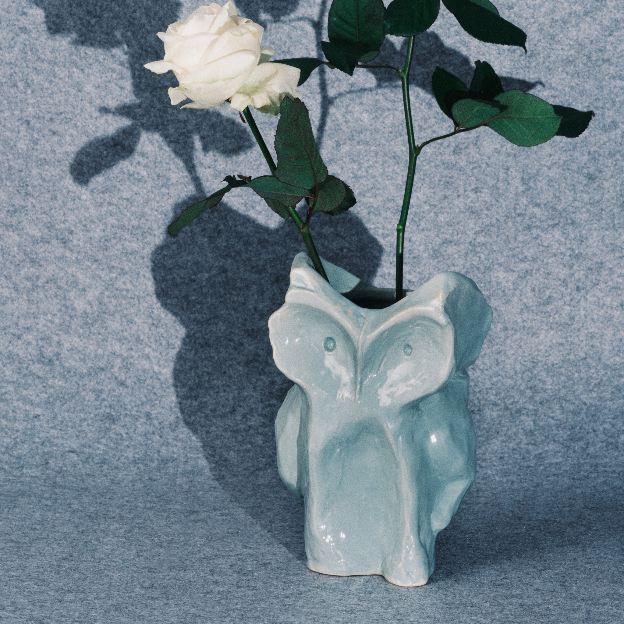 Een witte roos met groene bladeren zit in de Serax After Midnight Uil Vaas Lichtblauw S, een lichtblauwe keramische vaas in de vorm van een uil van Serax, tegen een grijs gestructureerde achtergrond.