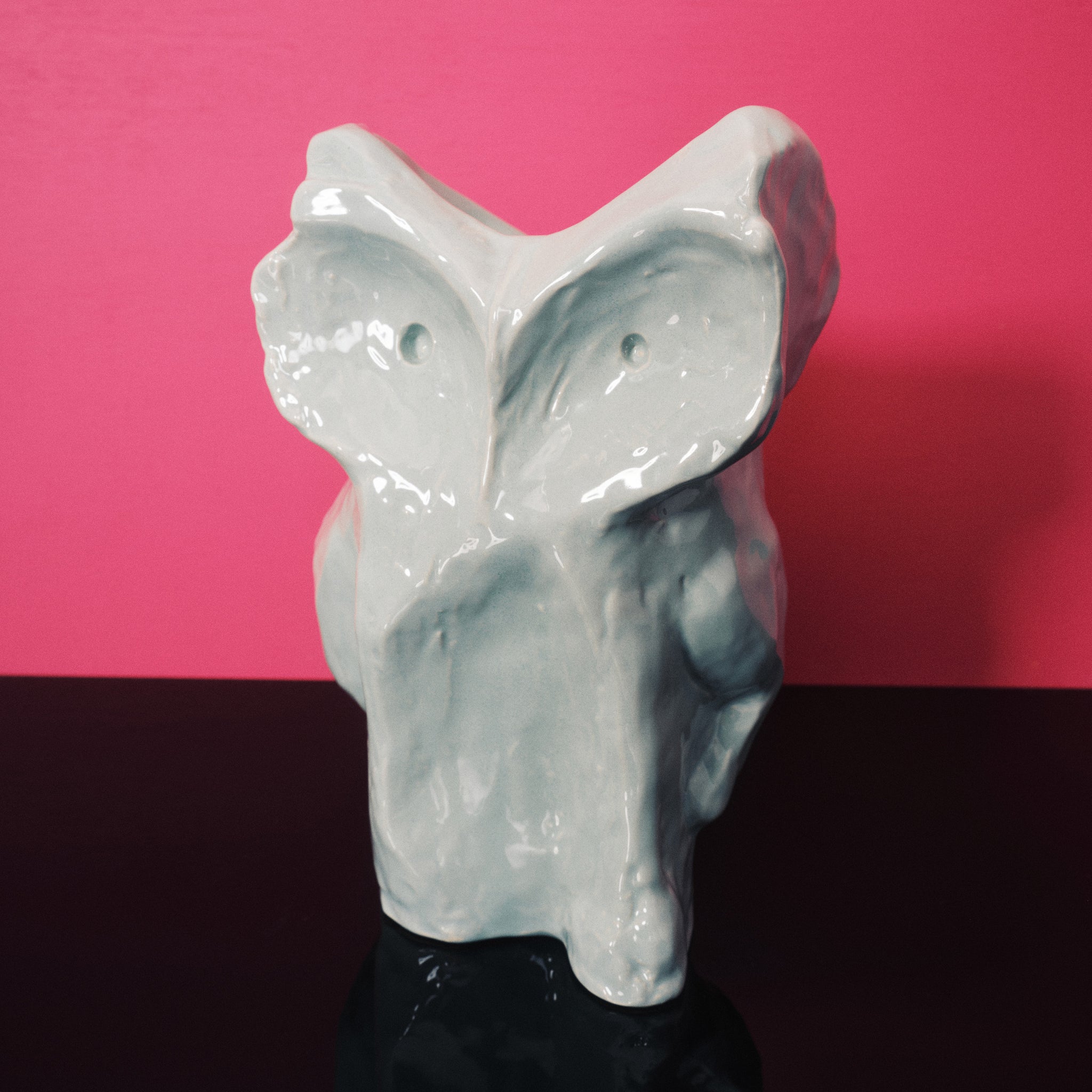De Serax After Midnight Uil Vaas Lichtblauw S, een abstracte glanzende keramische uilsculptuur met ronde vormen en kleine ooggaten, staat op een zwart oppervlak tegen een felroze achtergrond.