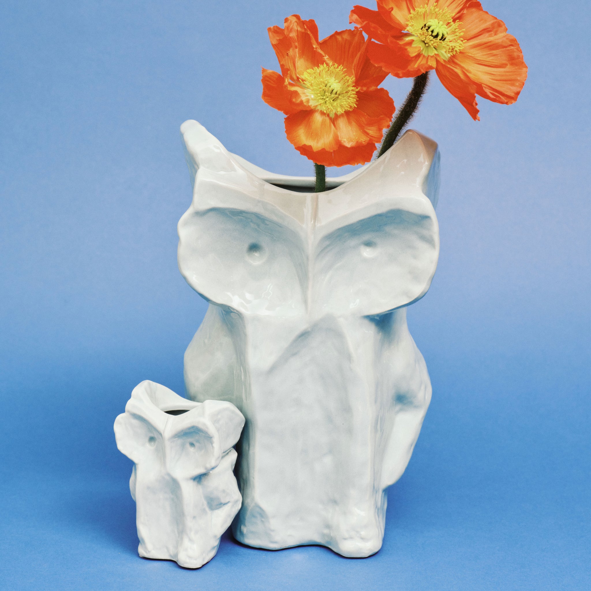 De Serax After Midnight Uil Vaas Lichtblauw S, een vaas in de vorm van een uil van wit keramiek, is afgebeeld op een blauwe achtergrond; in de grotere vaas zitten oranje klaprozen terwijl de kleinere vaas links ervan leeg is.