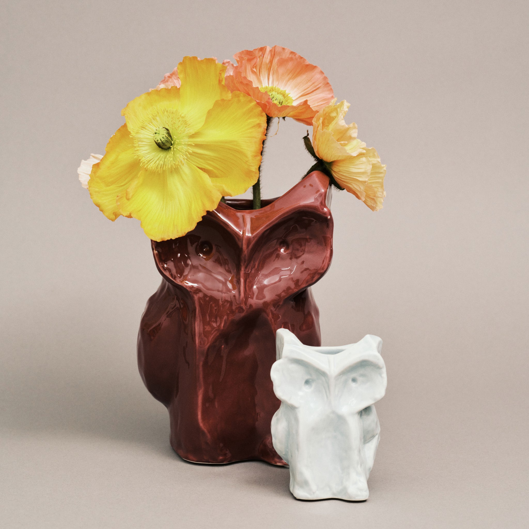 Een lichtblauwe Serax After Midnight Uil Vaas XS houdt gele en oranje klaproosbloemen vast achter een klein wit uilenfiguurtje van keramiek, beide tegen een effen beige achtergrond.