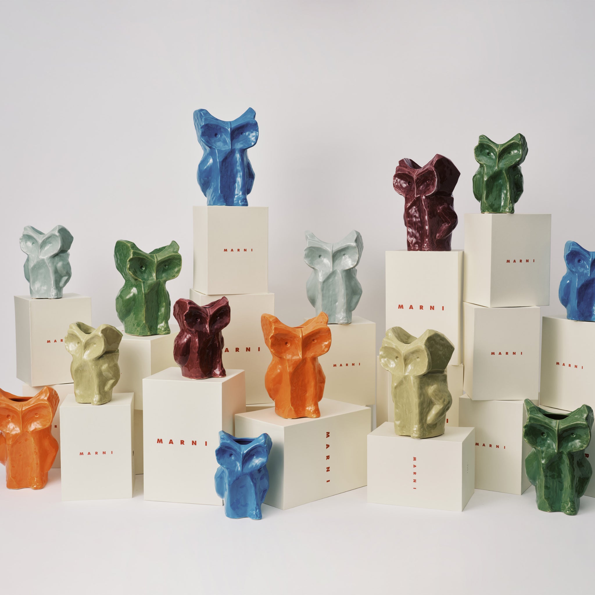 Kleurrijke abstracte keramische sculpturen, zoals de Serax After Midnight Uil Vaas Aubergine XS, staan op witte dozen op verschillende hoogtes tegen een effen witte achtergrond.